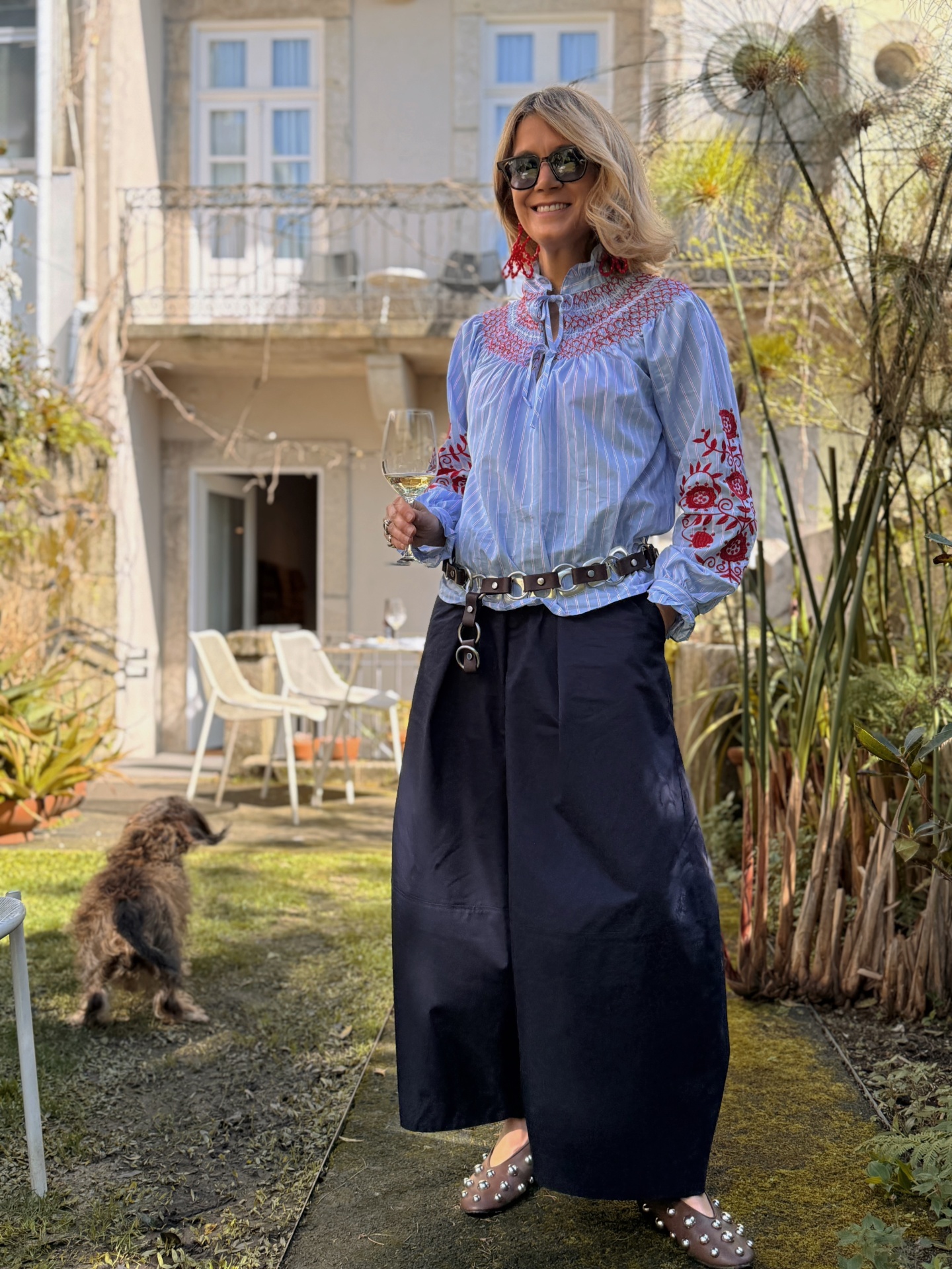 Pessoa com blusa azul clara com riscas brancas e bordados vermelhos, calças pretas largas, cinto de couro, sapatos com tachas, óculos escuros e brincos vermelhos, segurando copo de vinho