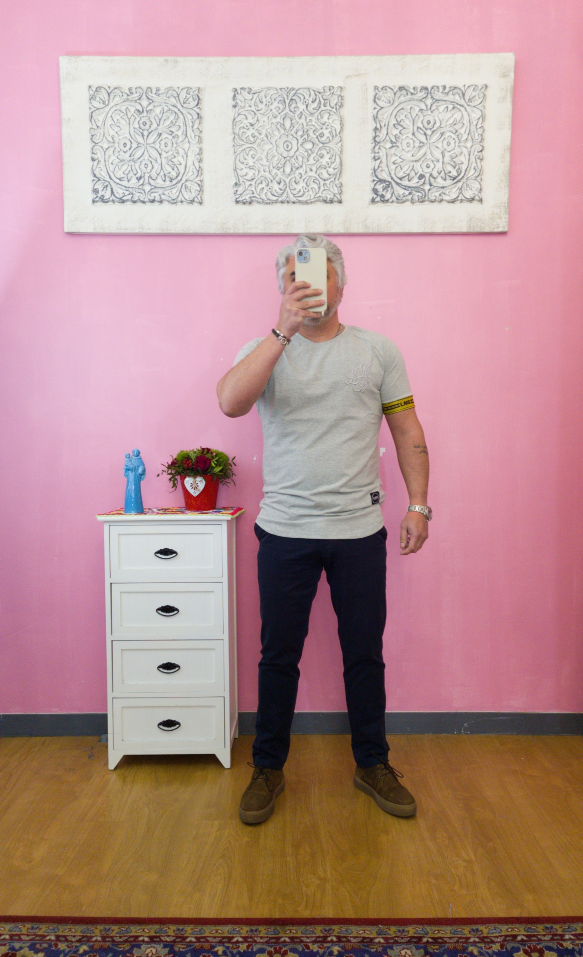 Homem com t-shirt cinza e calças escuras a tirar foto em quarto com parede rosa e móvel branco