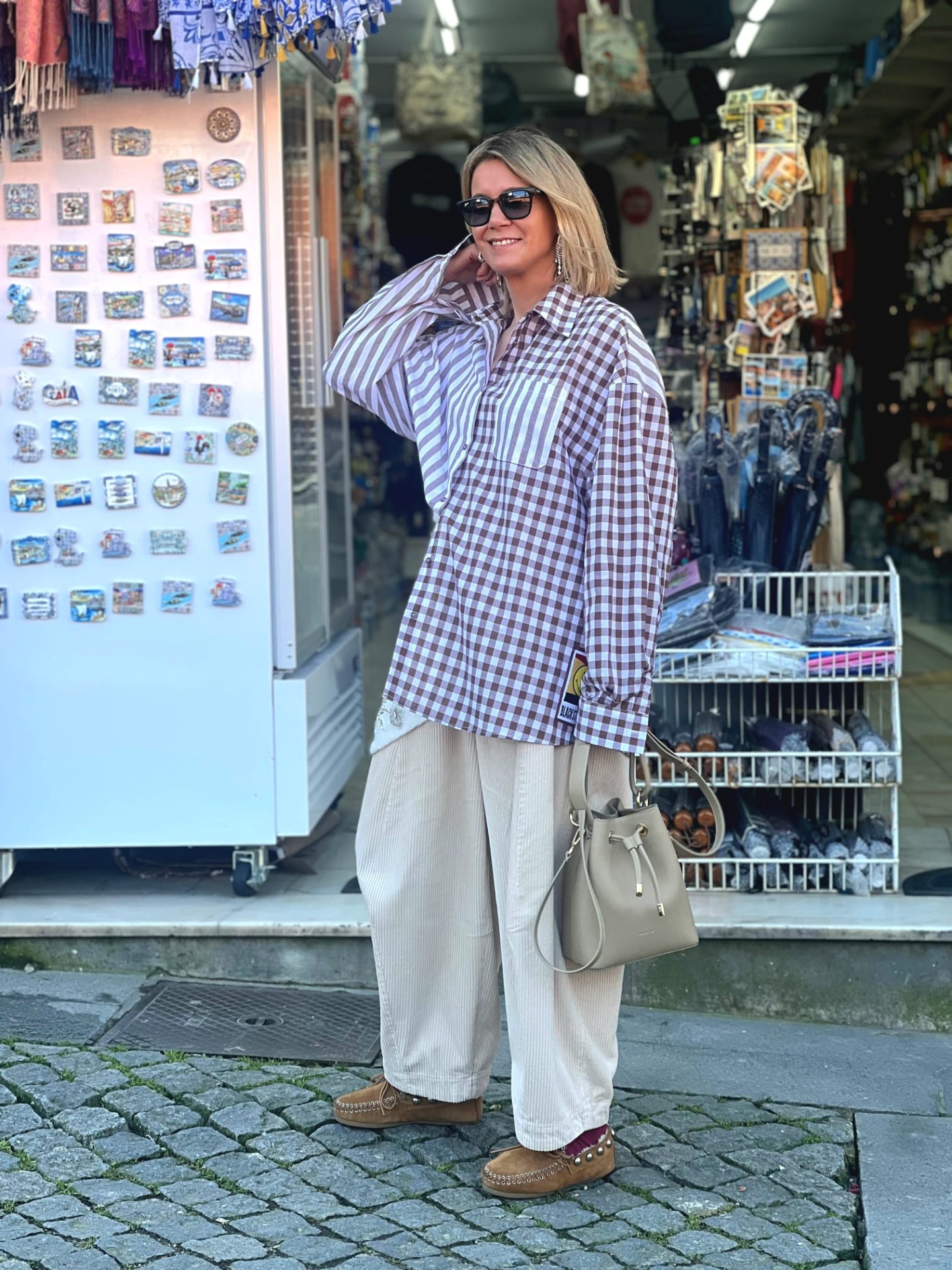 Mulher com camisa xadrez castanha e branca, calças creme largas, mocassins e mala bege na rua com lojas ao fundo
