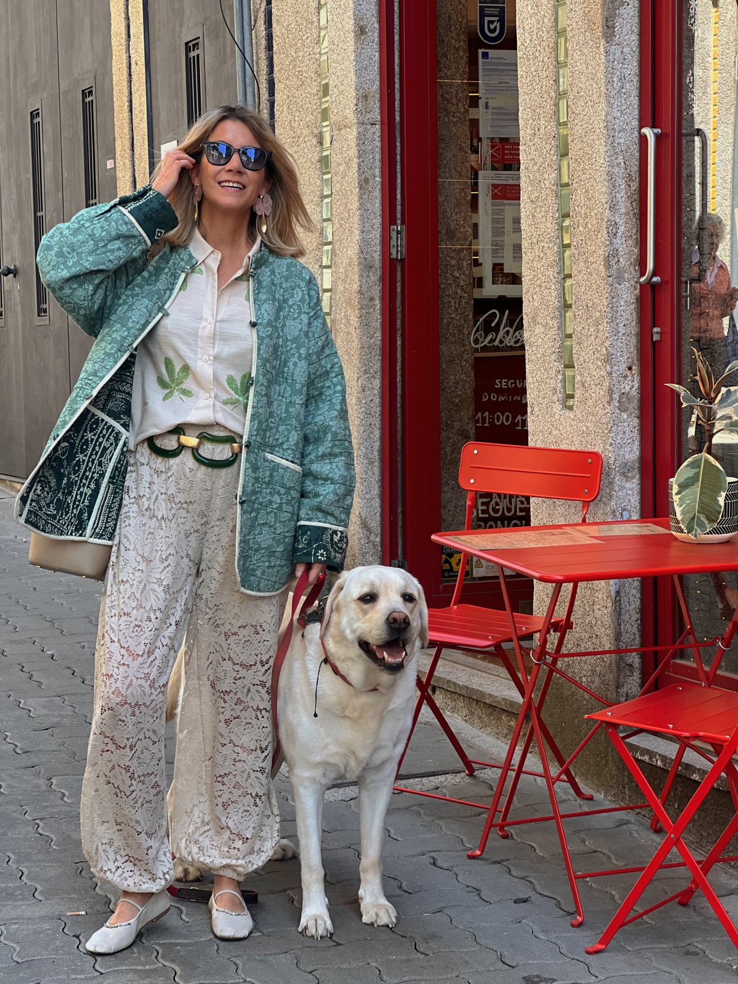 Mulher com casaco verde e calças rendadas na rua com cão branco ao lado