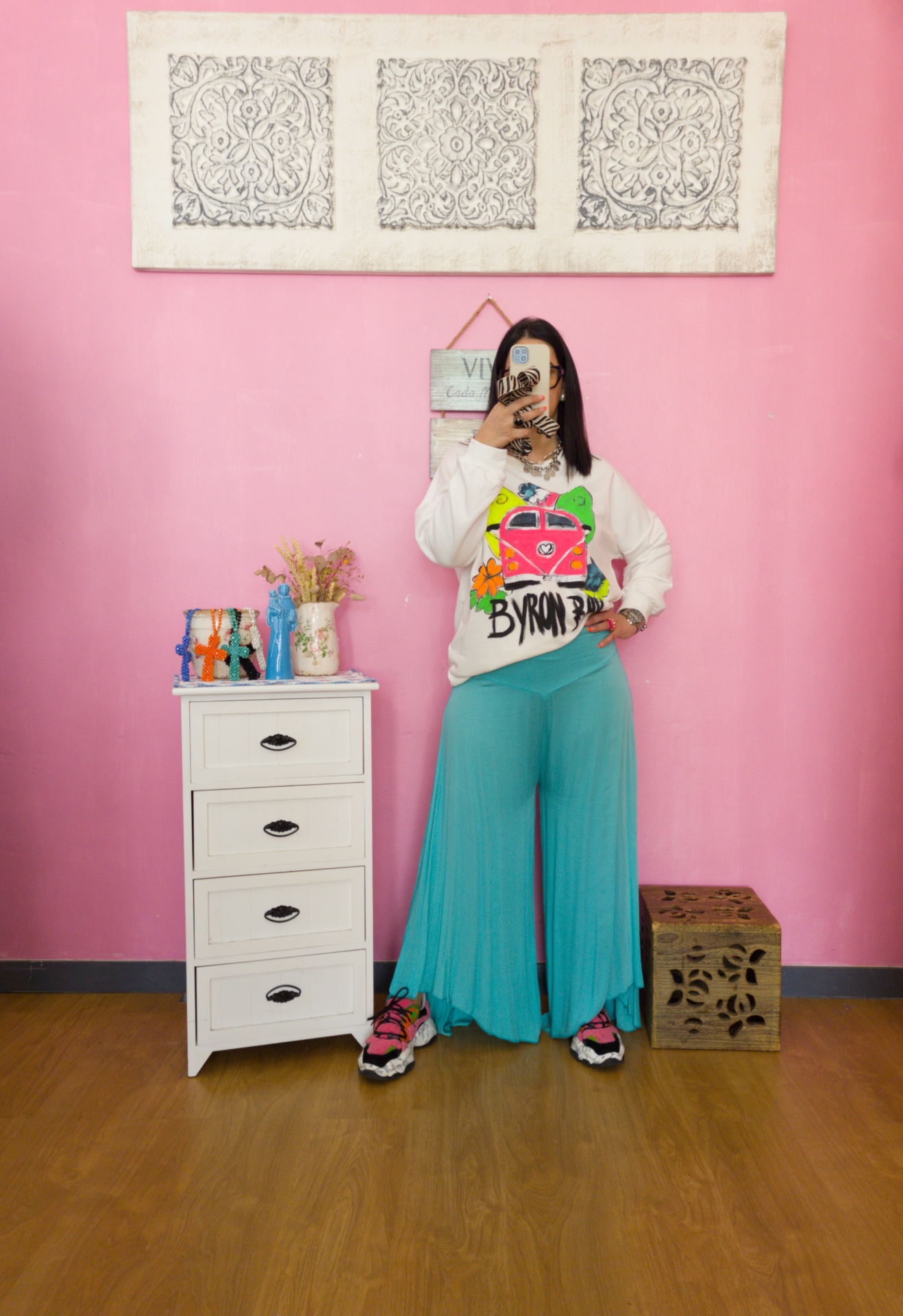 Pessoa a usar camisola branca estampada e calças largas azuis, de pé contra parede rosa com móvel branco ao lado