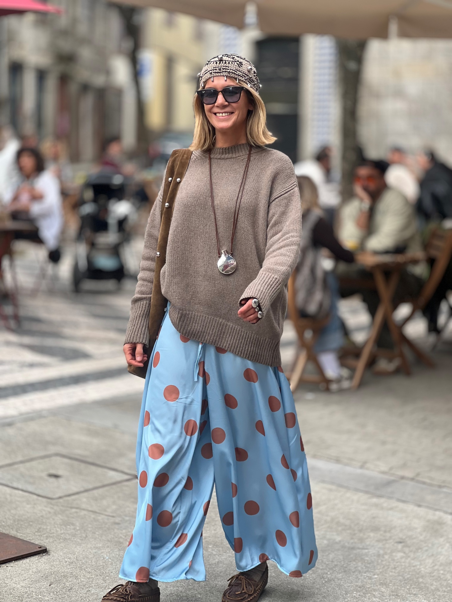 Mulher com camisola castanha, calças azuis com bolinhas castanhas e lenço na cabeça numa rua com esplanada.