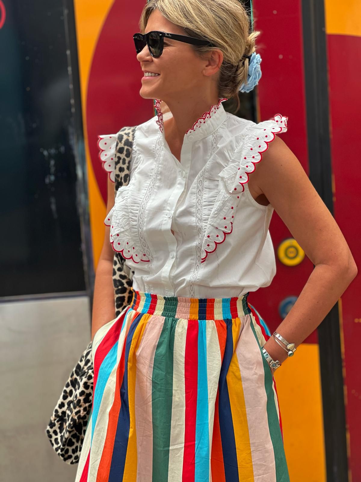 Mulher com blusa branca bordada, saia listrada colorida e óculos escuros em fundo colorido