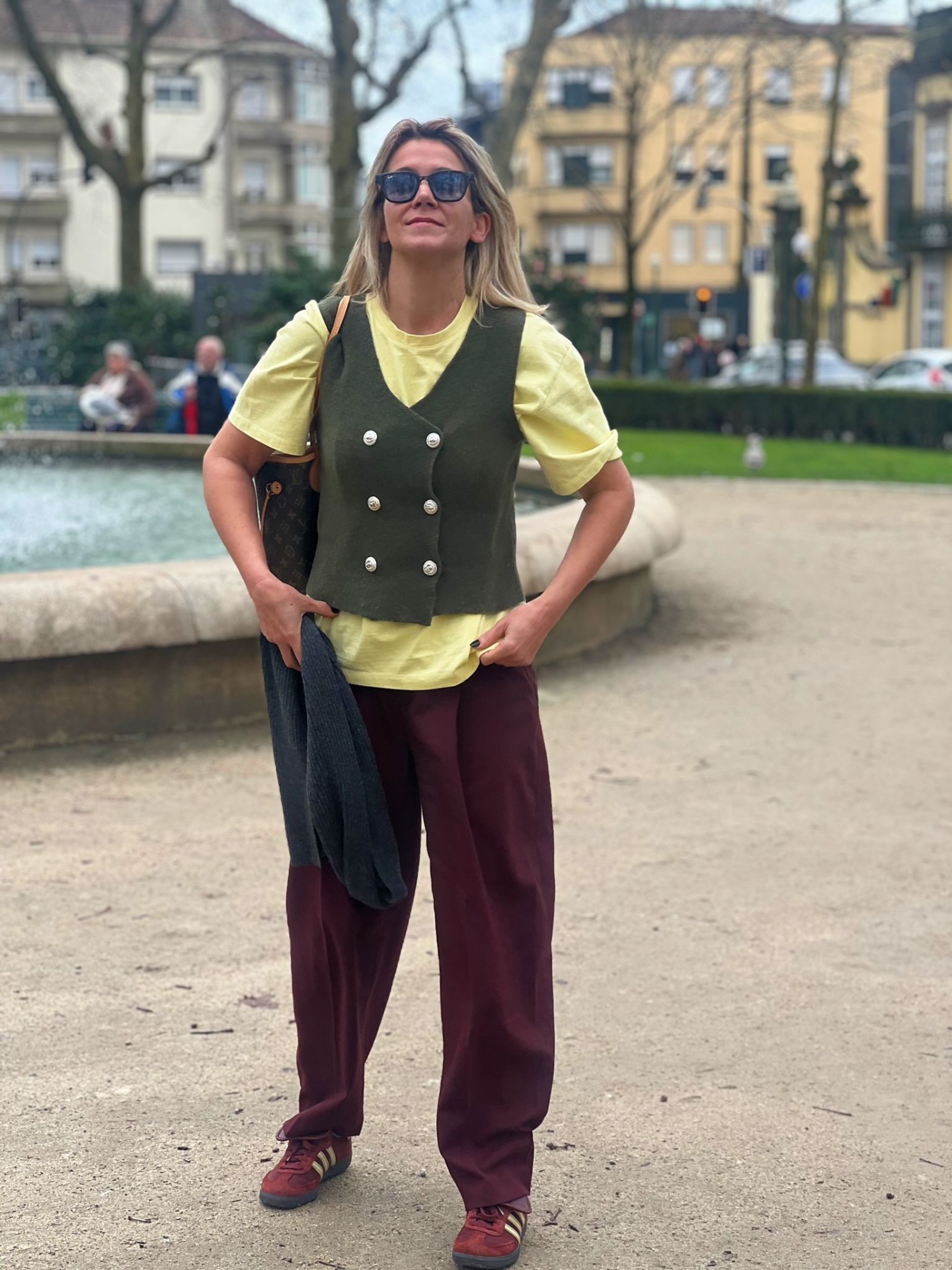 Mulher com colete verde-oliva, t-shirt amarela, calças vinho e ténis vermelhos num parque