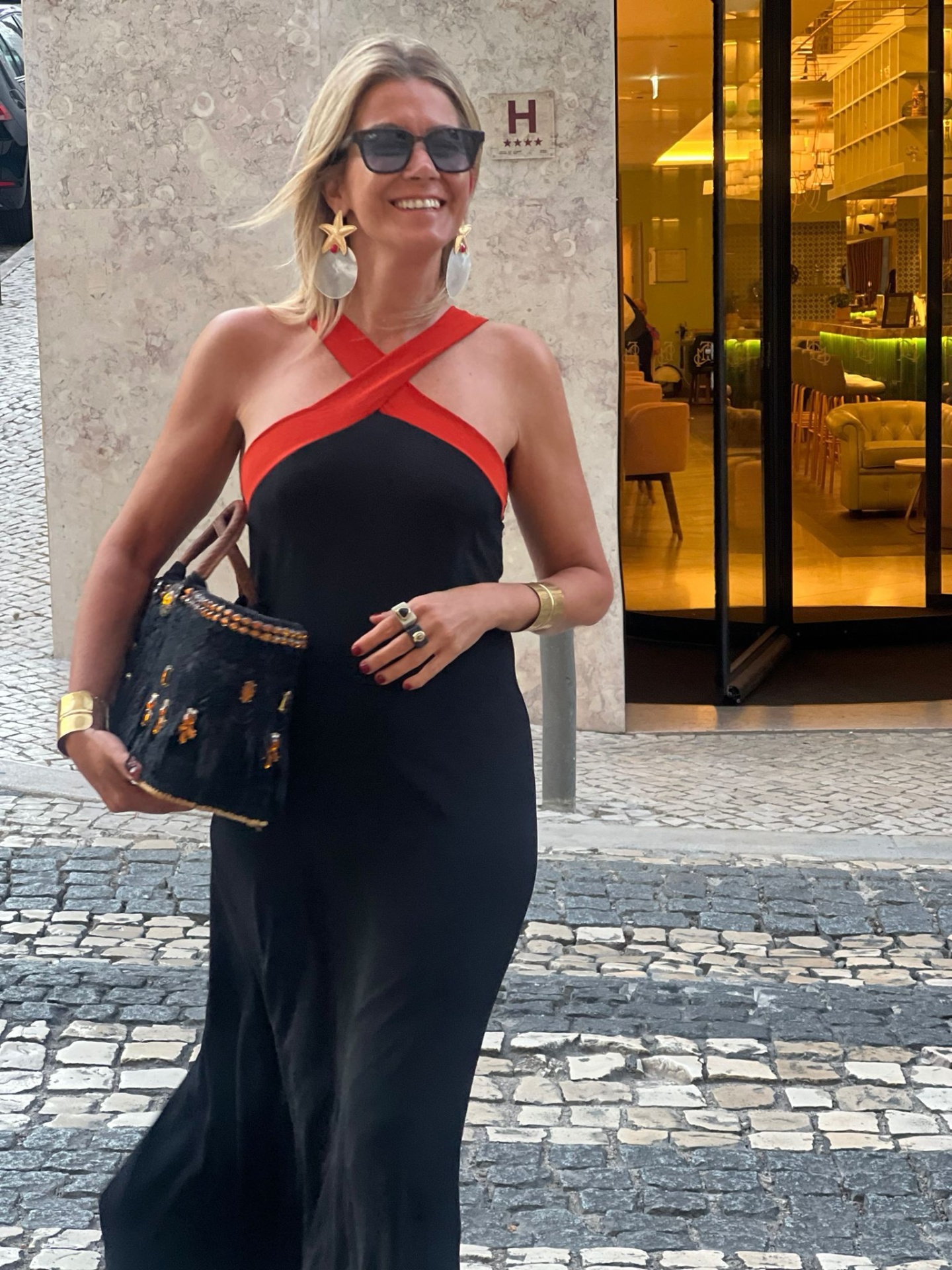 Mulher com vestido longo preto e vermelho, acessórios dourados e mala preta na calçada