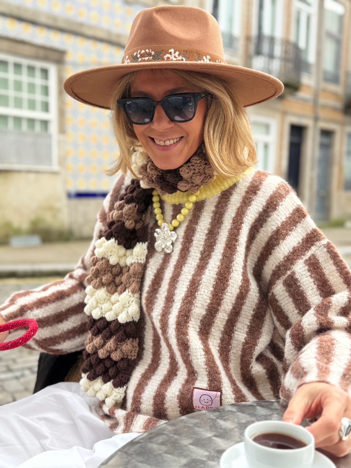 Mulher com casaco riscado, echarpe castanha, chapéu castanho e óculos escuros a beber chá