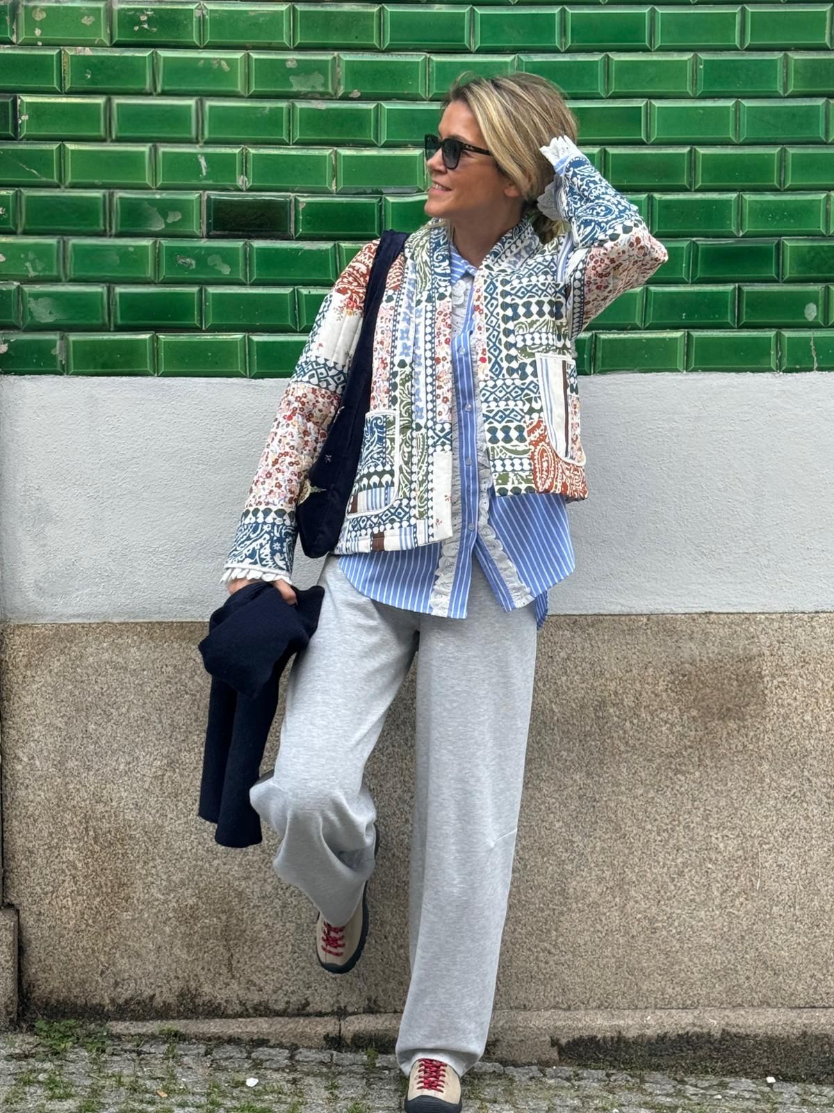 Pessoa com casaco patchwork colorido e óculos em frente a parede de azulejos verdes