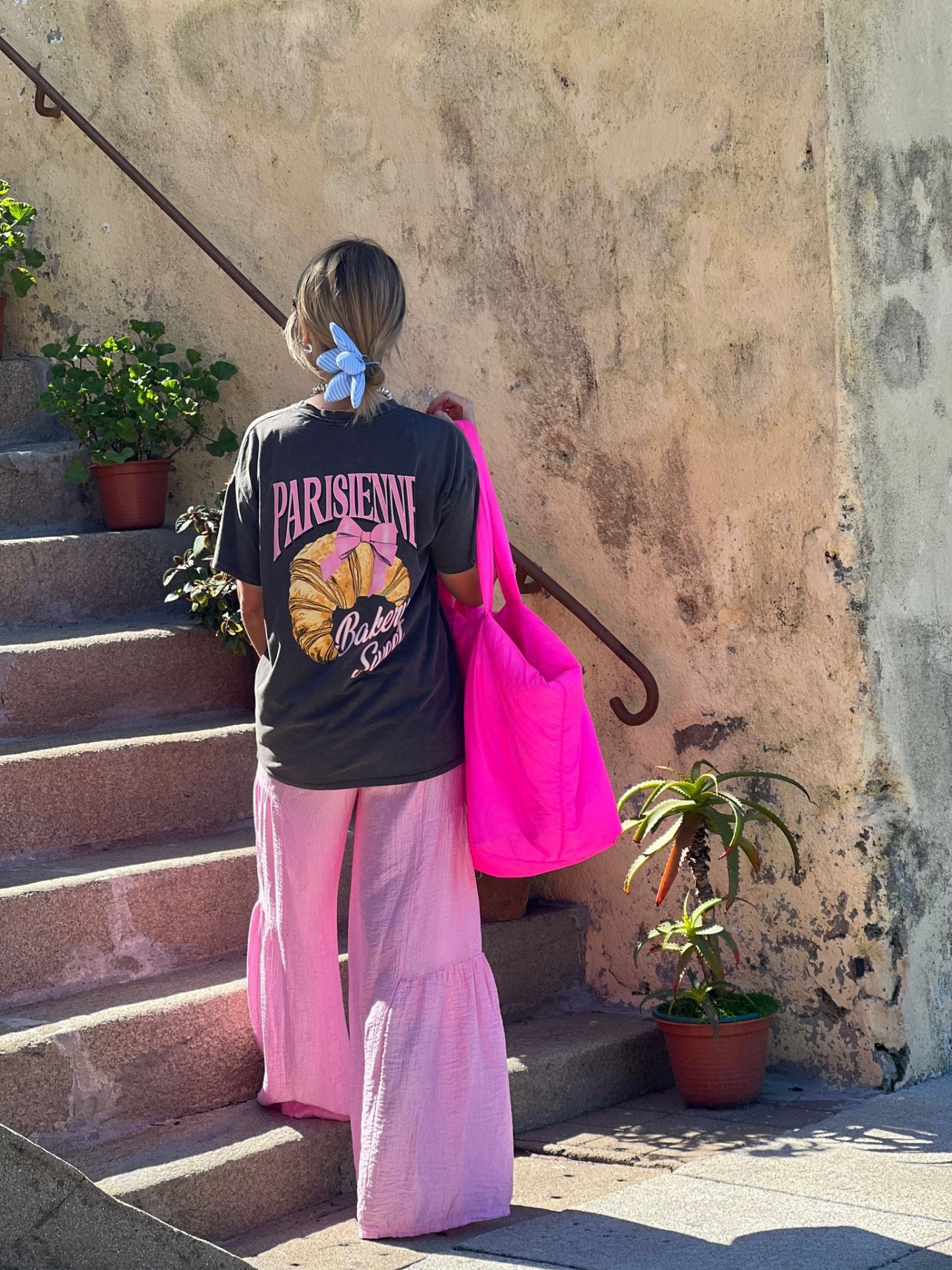 Pessoa de costas com roupa casual e bolsa rosa subindo escadas externas com parede envelhecida e plantas.