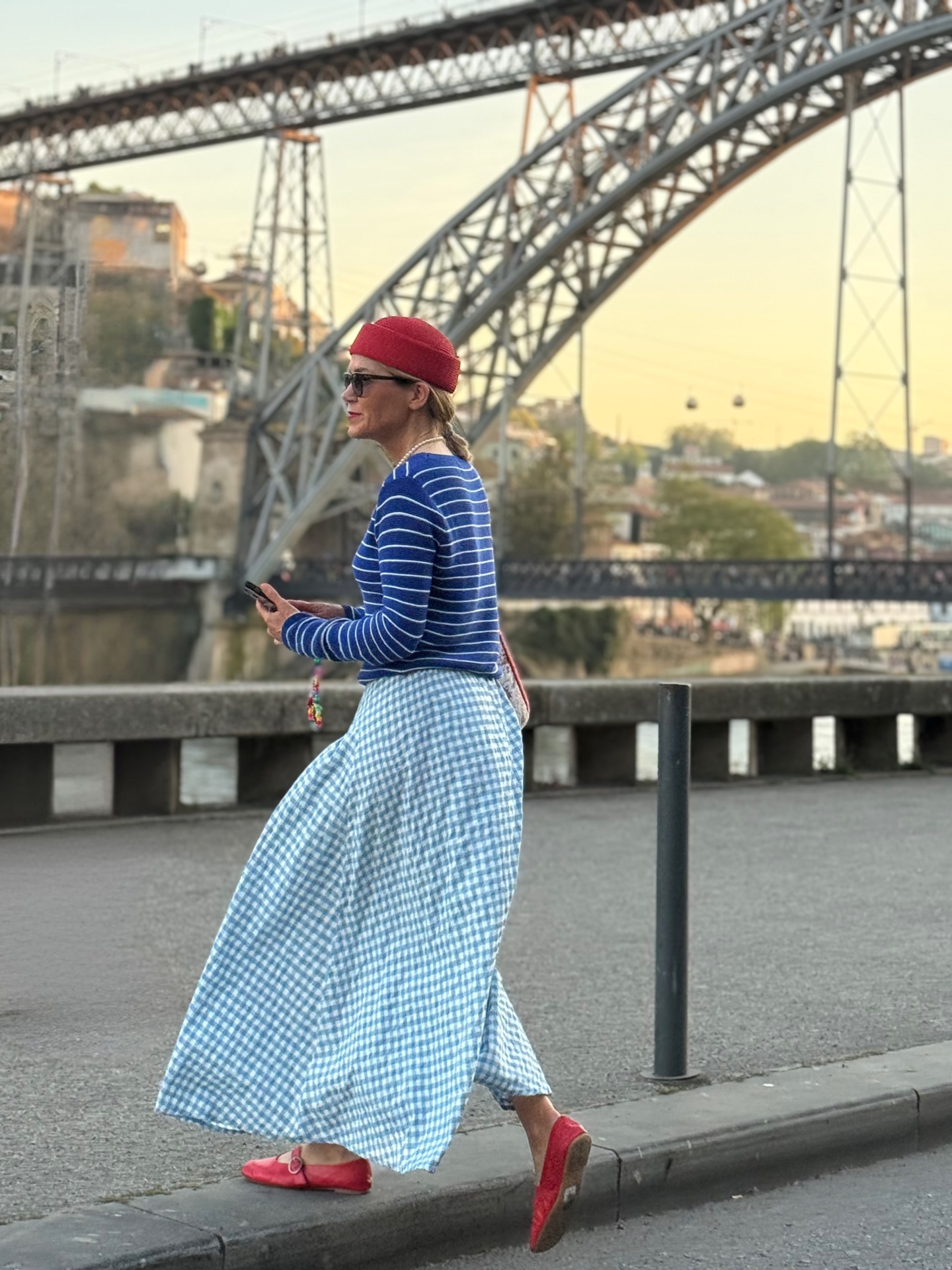 Mulher com camisola azul às riscas brancas, saia azul e branca padrão vichy, sapatos vermelhos e chapéu vermelho numa avenida junto a ponte metálica.