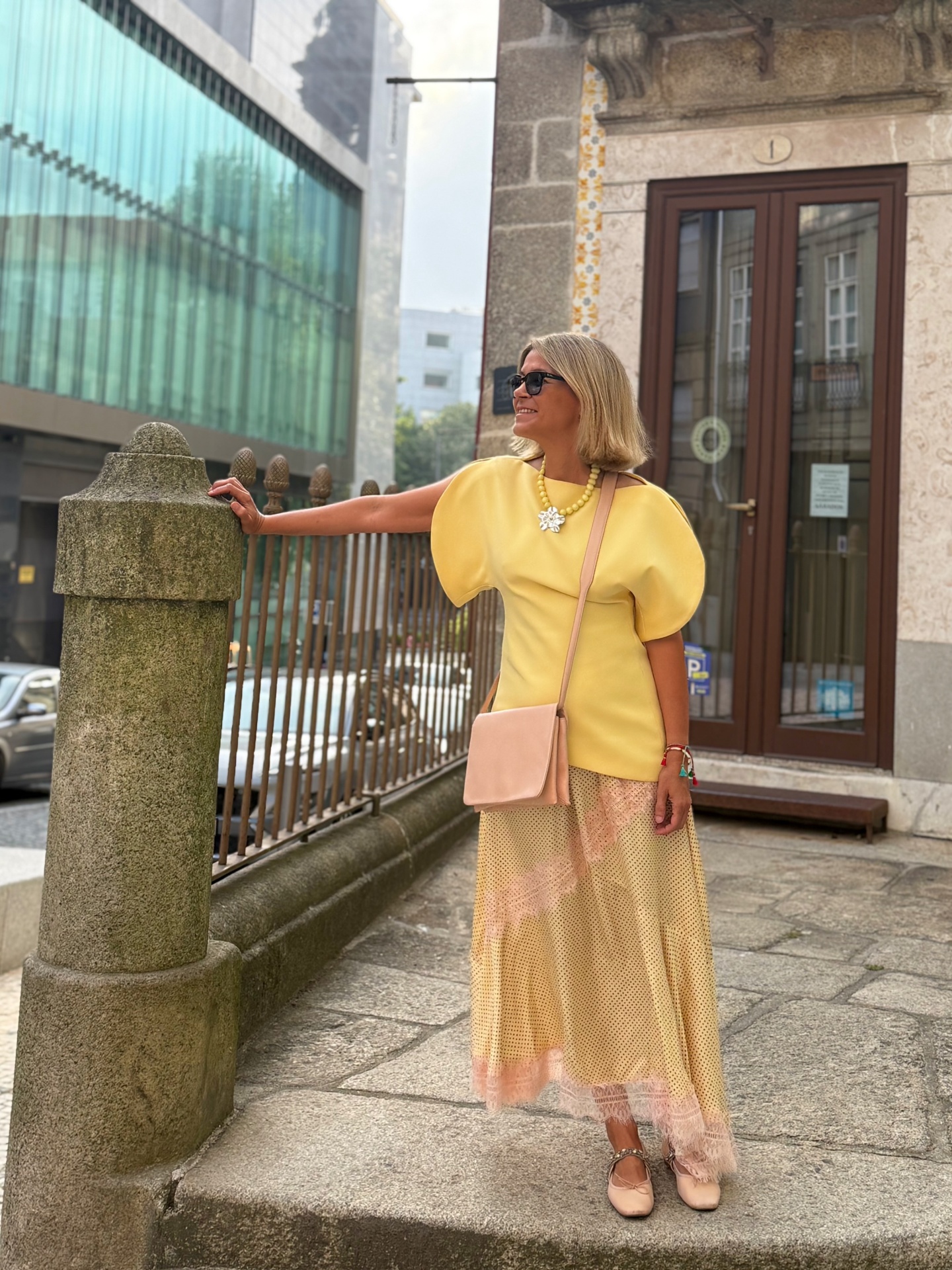 Mulher de vestido amarelo e saia comprida bege com acessórios coloridos em ambiente urbano.