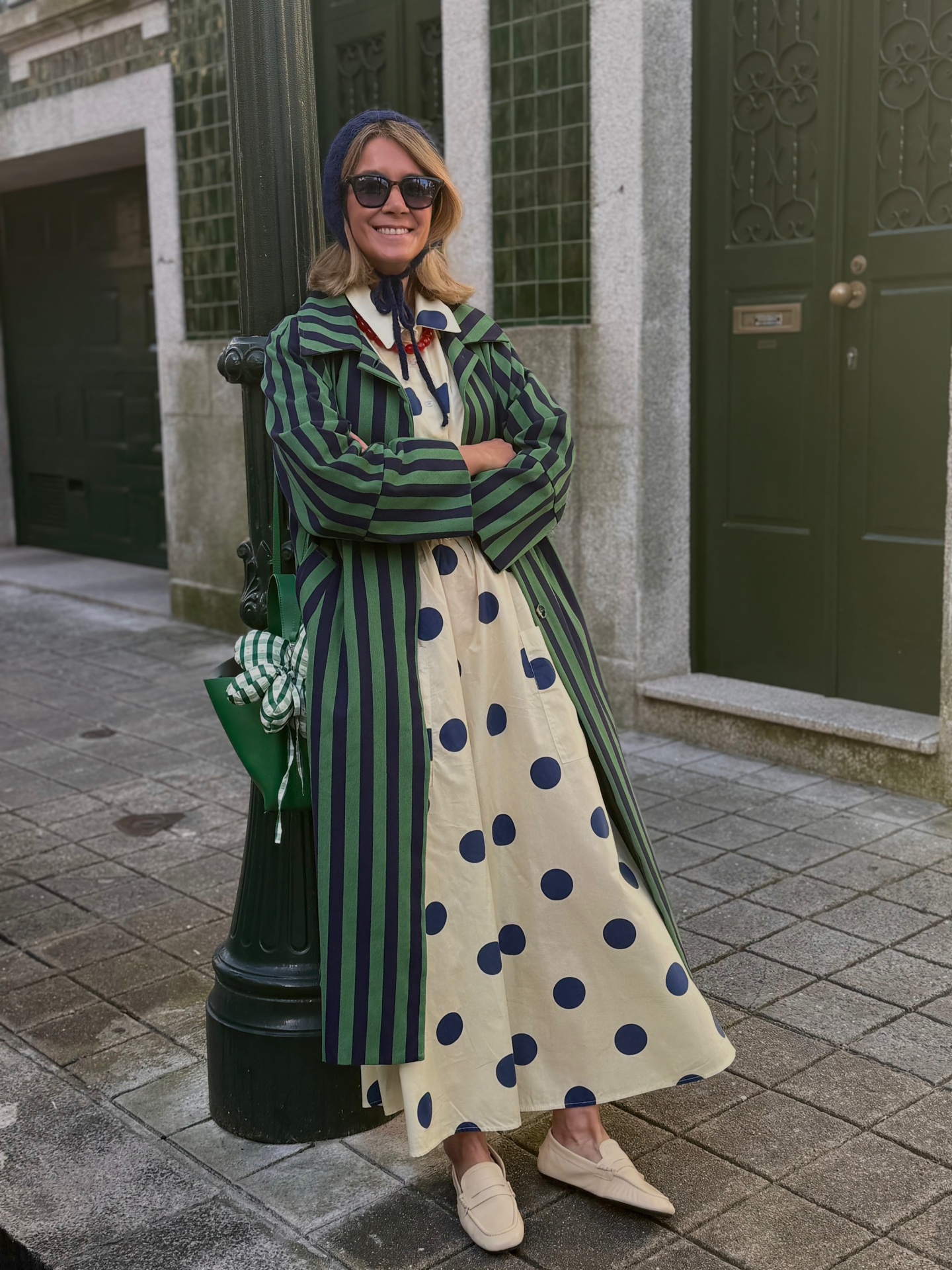 Mulher com casaco riscado verde e preto, vestido creme com bolinhas azuis, sapatos bege, saco verde com lenço, gorro azul e óculos escuros