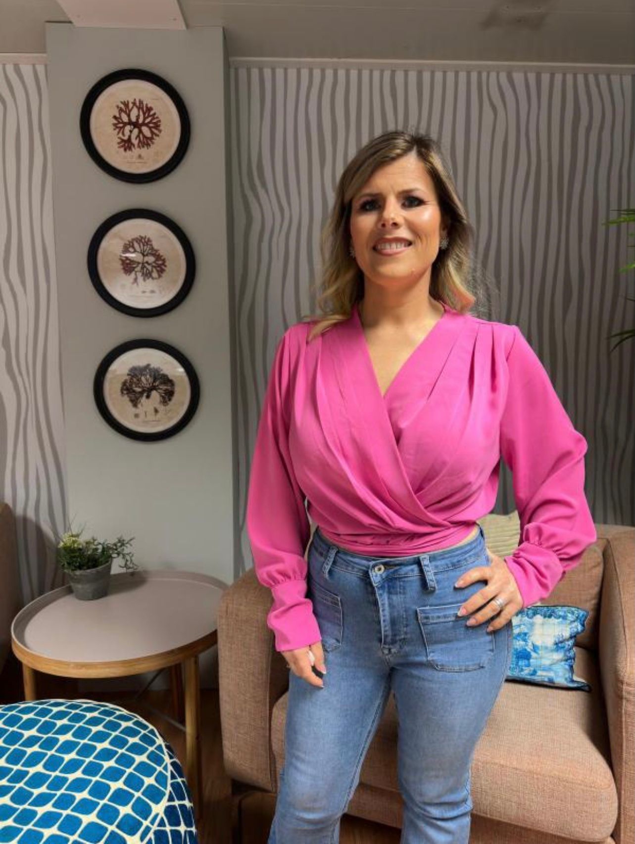 Mulher com blusa rosa e calças de ganga azuis em sala decorada com sofá, planta e quadros arbóreos