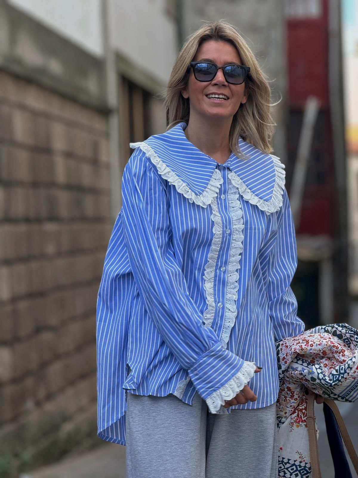 Mulher com camisa azul riscada branca e detalhes em renda, calças cinza, óculos escuros, segurando casaco floral numa rua