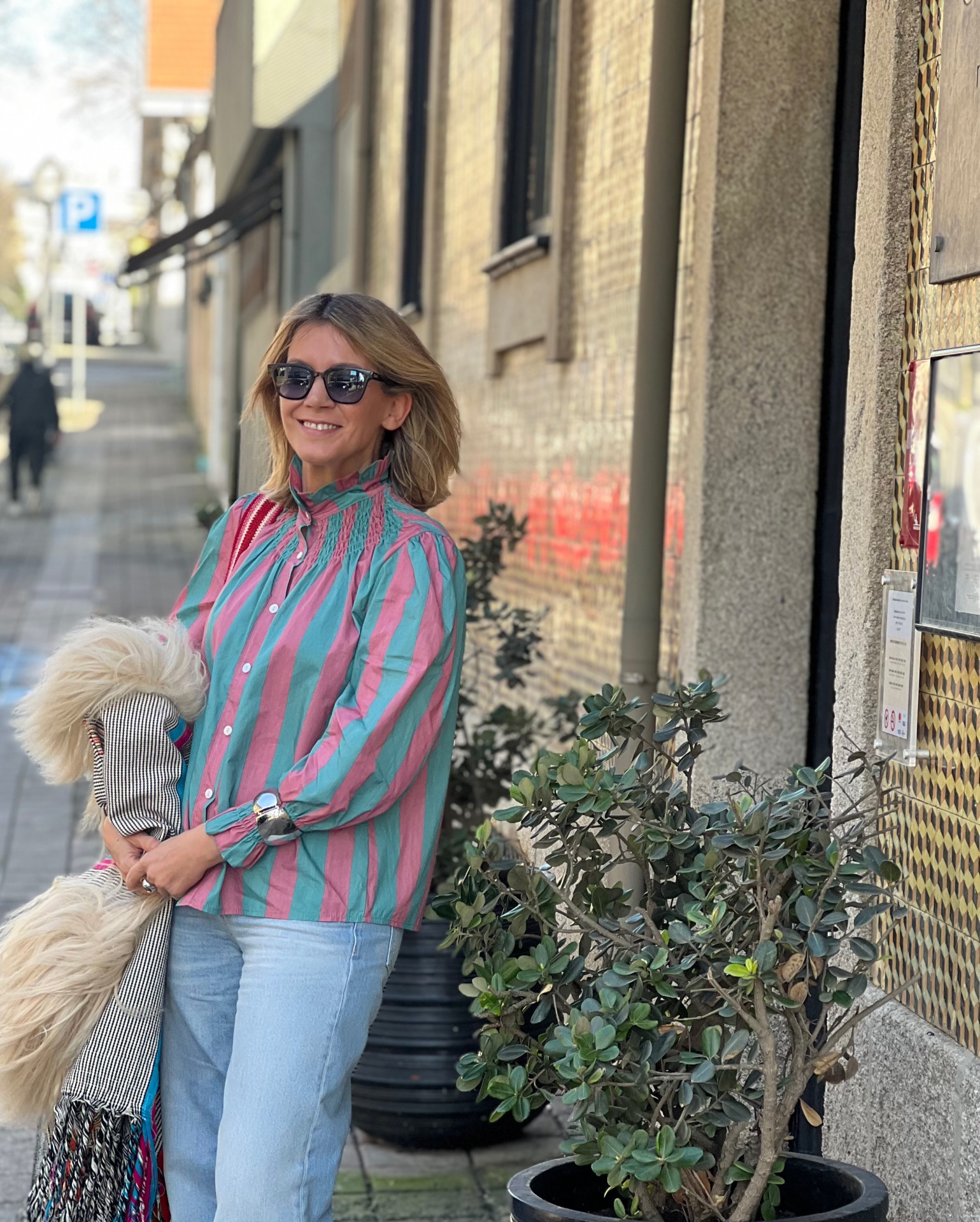 Blusa riscada rosa e verde com gola alta e punhos franzidos, jeans azuis claros, mulher com óculos escuros segurando casaco pêlo