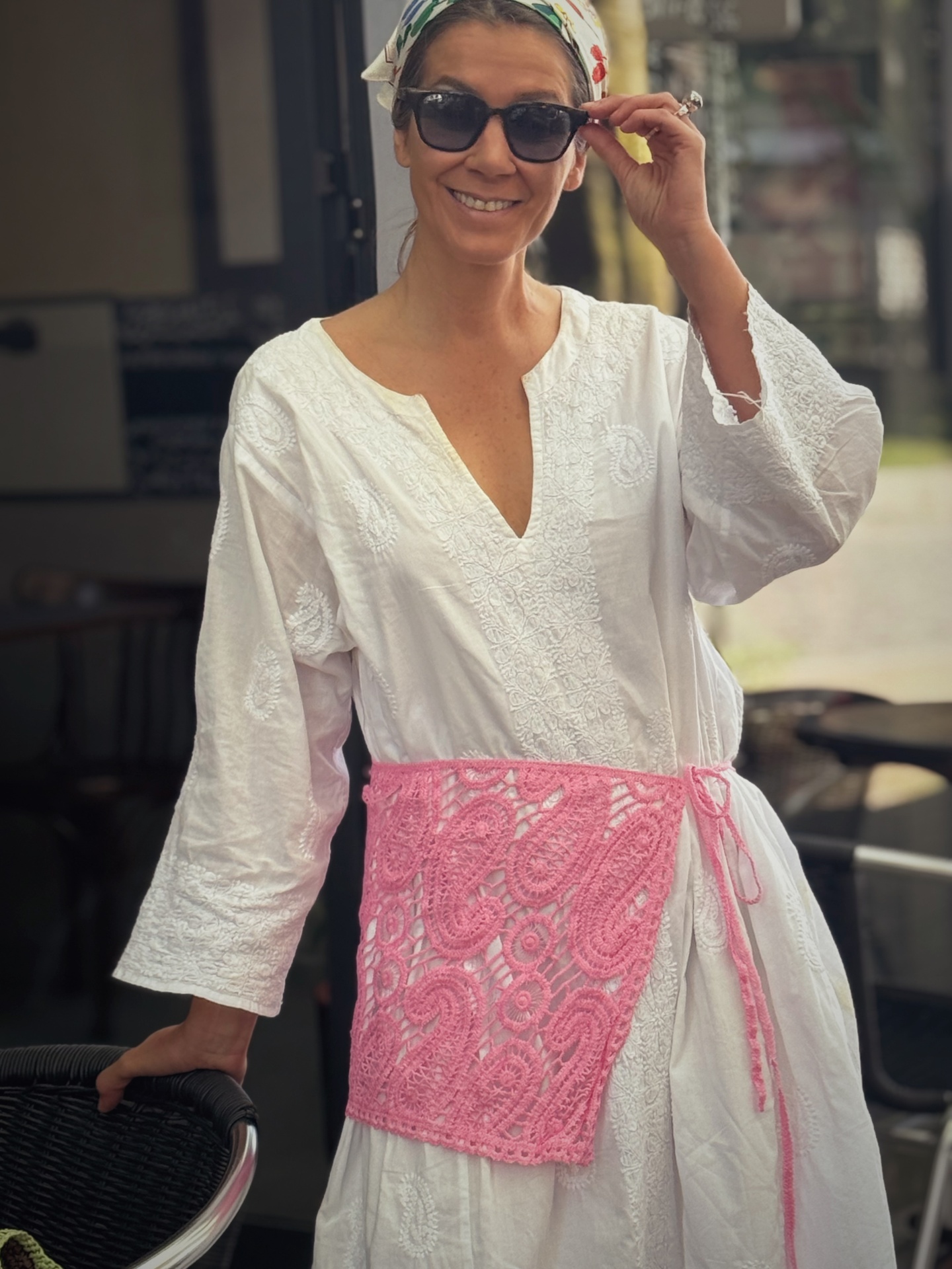 Vestido branco bordado, cinto rosa croché, óculos de sol pretos, lenço cabeça branco com padrão colorido.