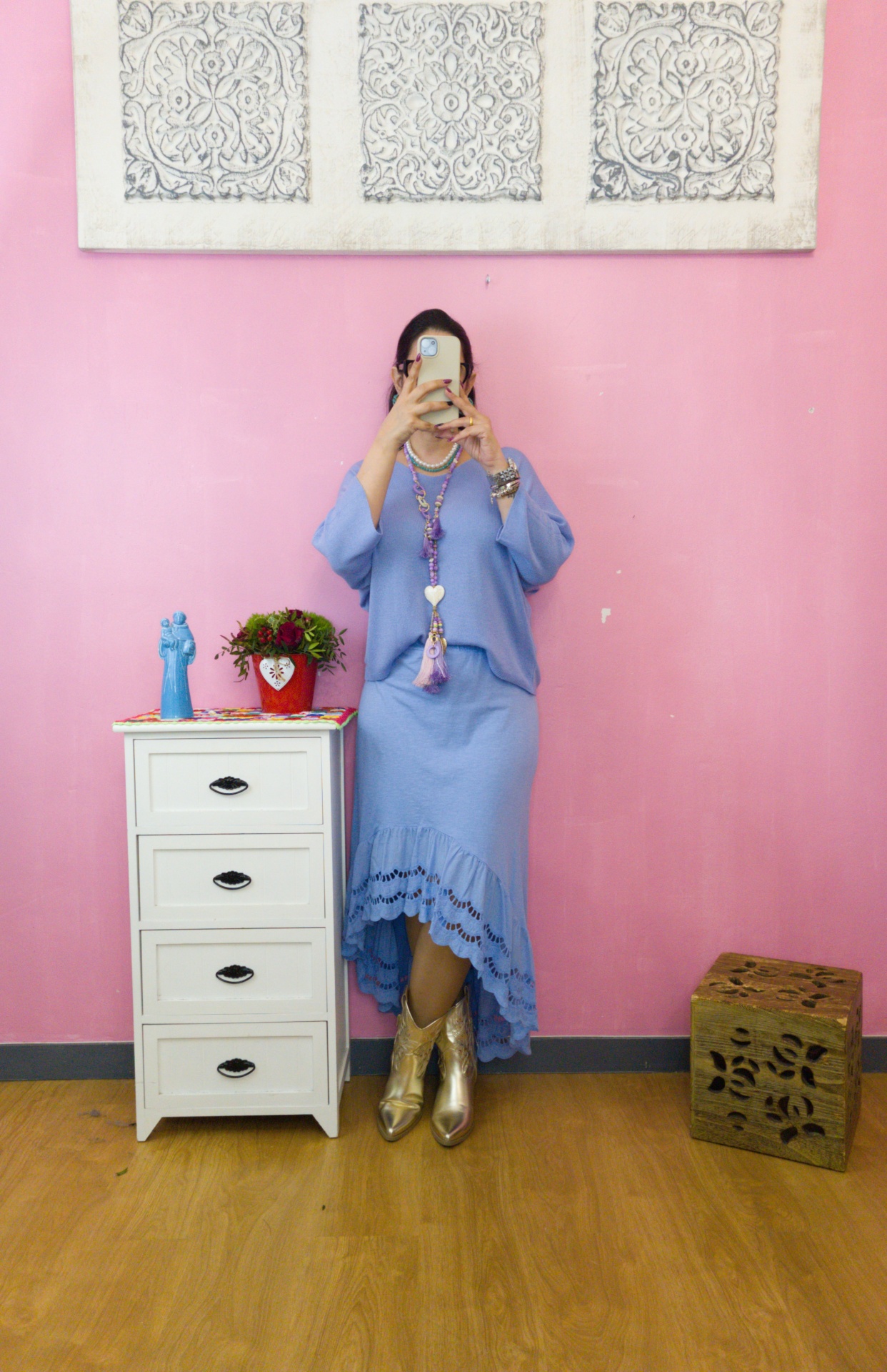 Pessoa com vestido azul claro e botas douradas encostada em cómoda branca com decoração, parede rosa e quadros decorativos.