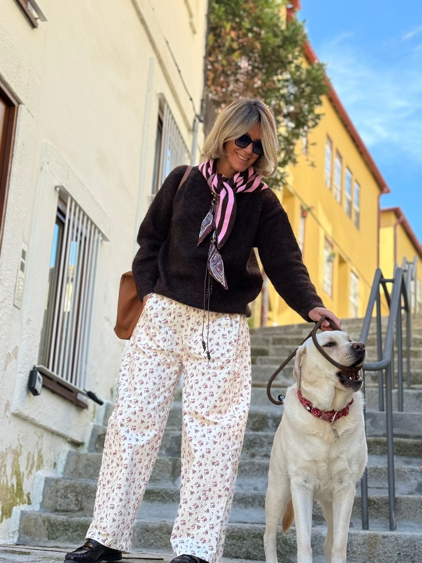 Mulher com calças largas brancas com padrão floral e casaco preto com cão Labrador branco numa escadaria