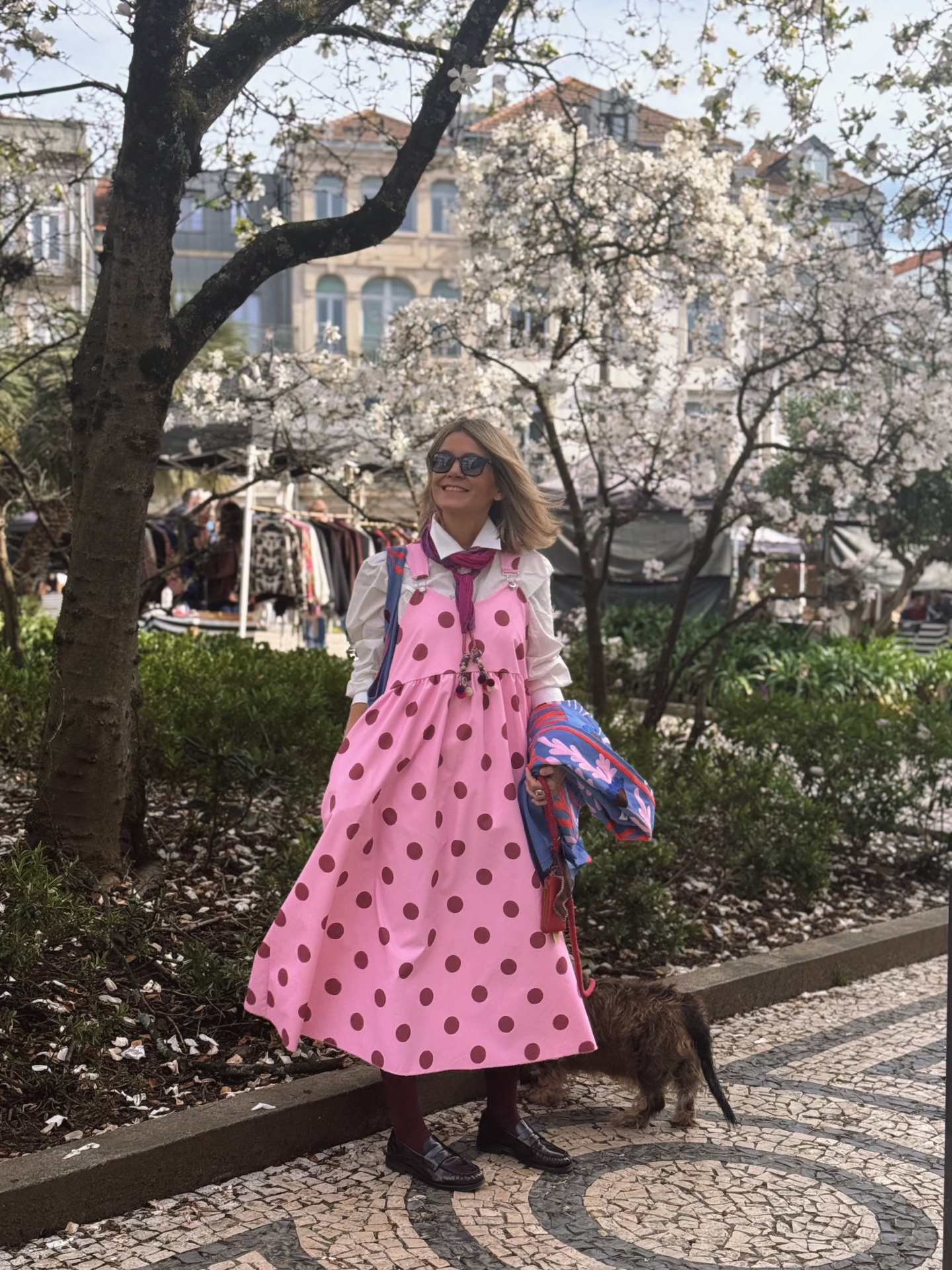 Mulher com vestido rosa às bolinhas castanhas na rua à sombra de árvores floridas com cão e calçada de mosaico.
