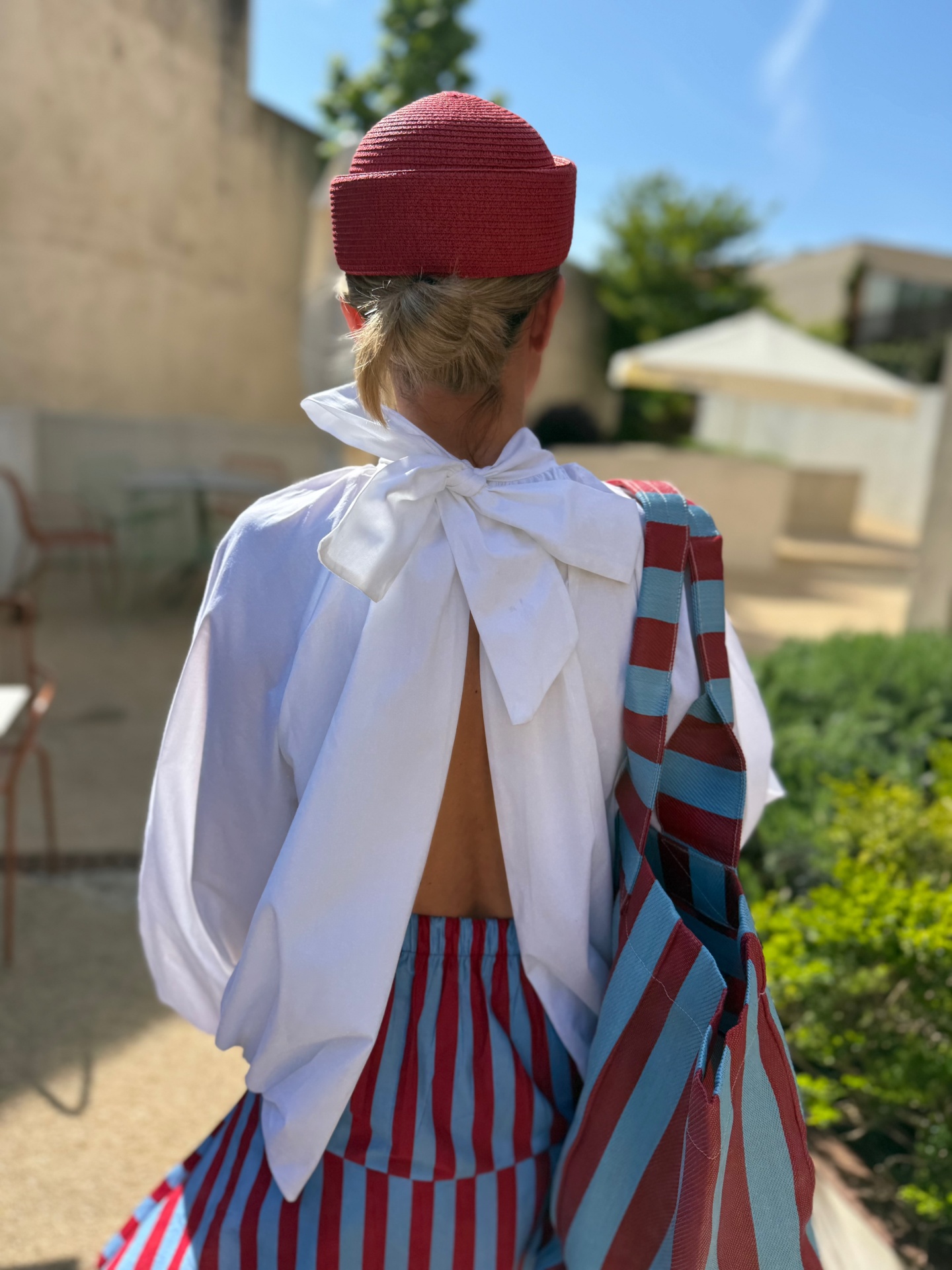 Pessoa com camisa branca aberta nas costas e saia riscada vermelha e azul, chapéu vermelho e bolsa riscada vermelha e azul, vista de costas