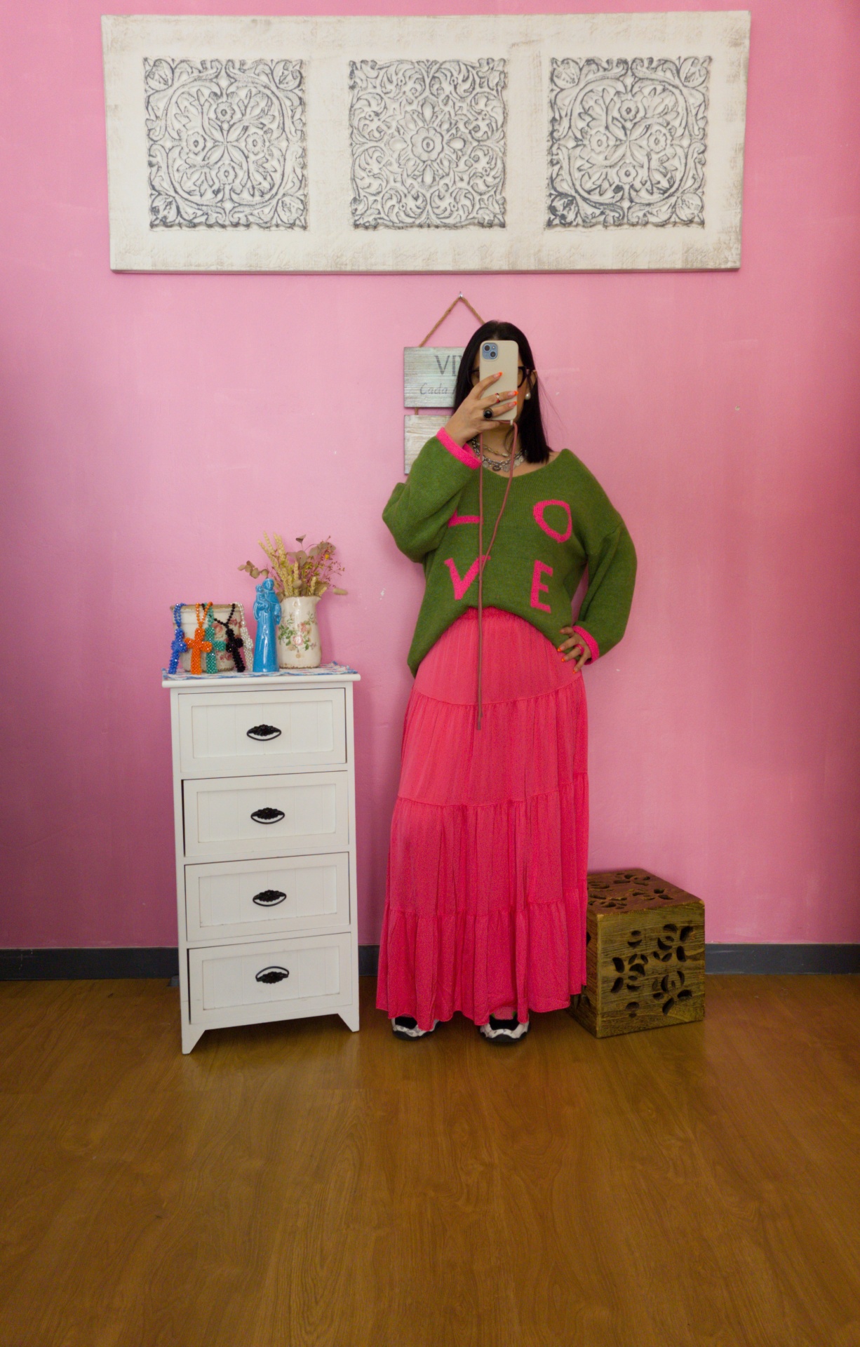 Mulher a tirar selfie com suéter verde e saia rosa em quarto com parede e móveis decorativos