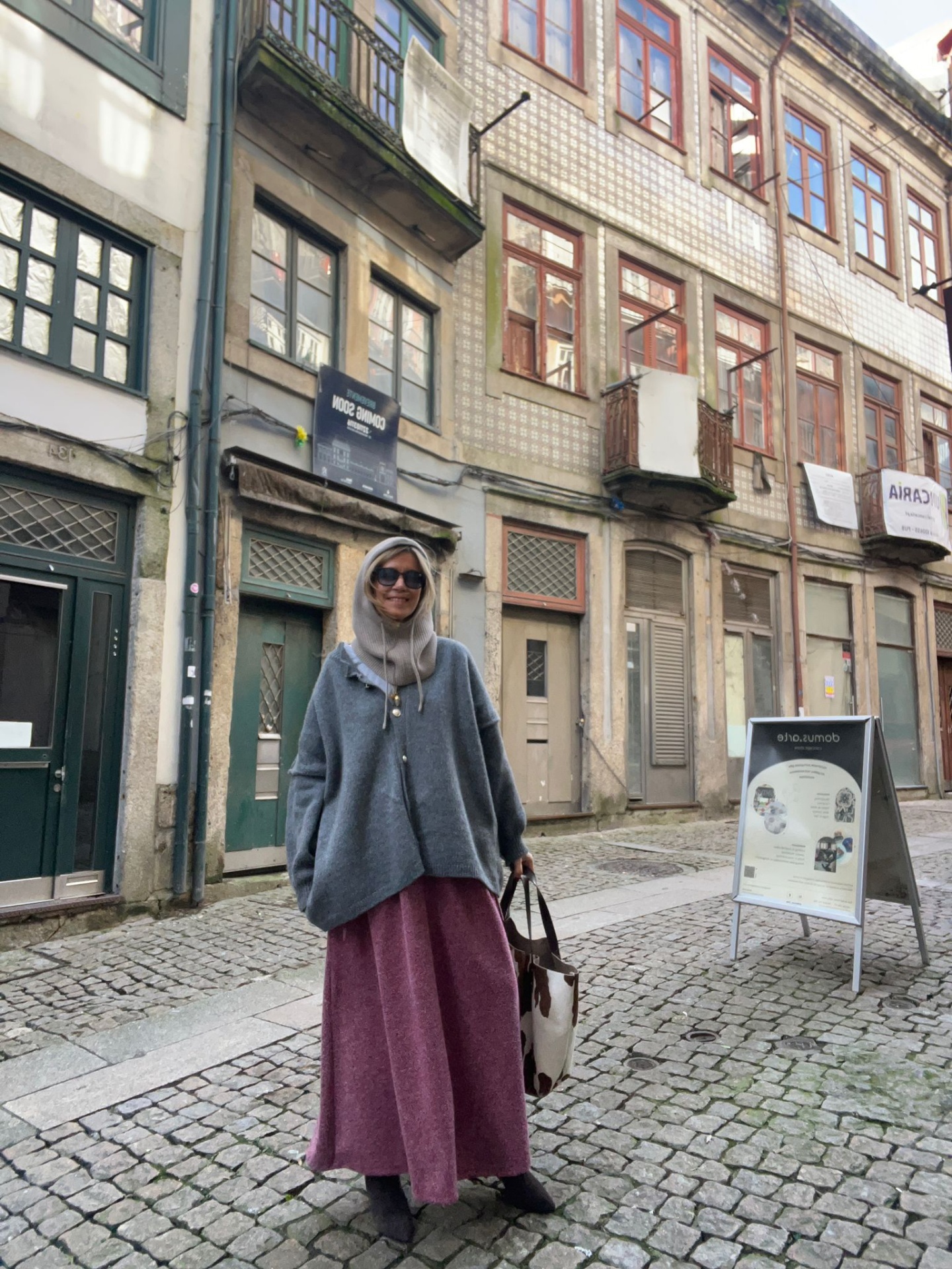 Mulher com poncho cinza e saia longa rosa numa rua de calçada com edifícios antigos ao fundo