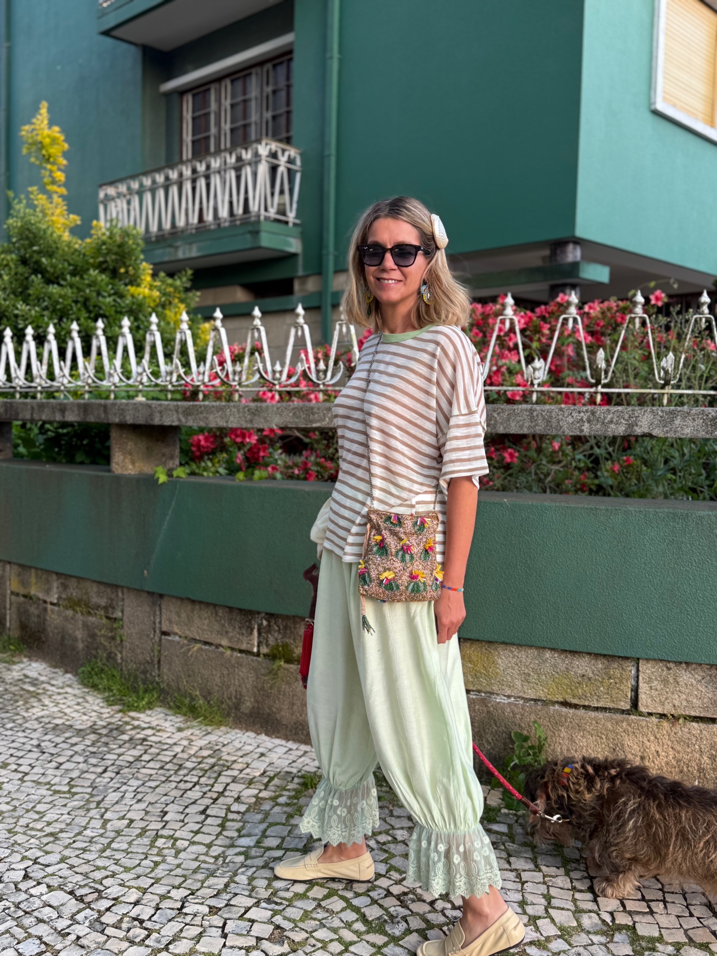 Mulher com calças largas rendadas, camisola às riscas, carteira floral e óculos de sol num passeio urbano.