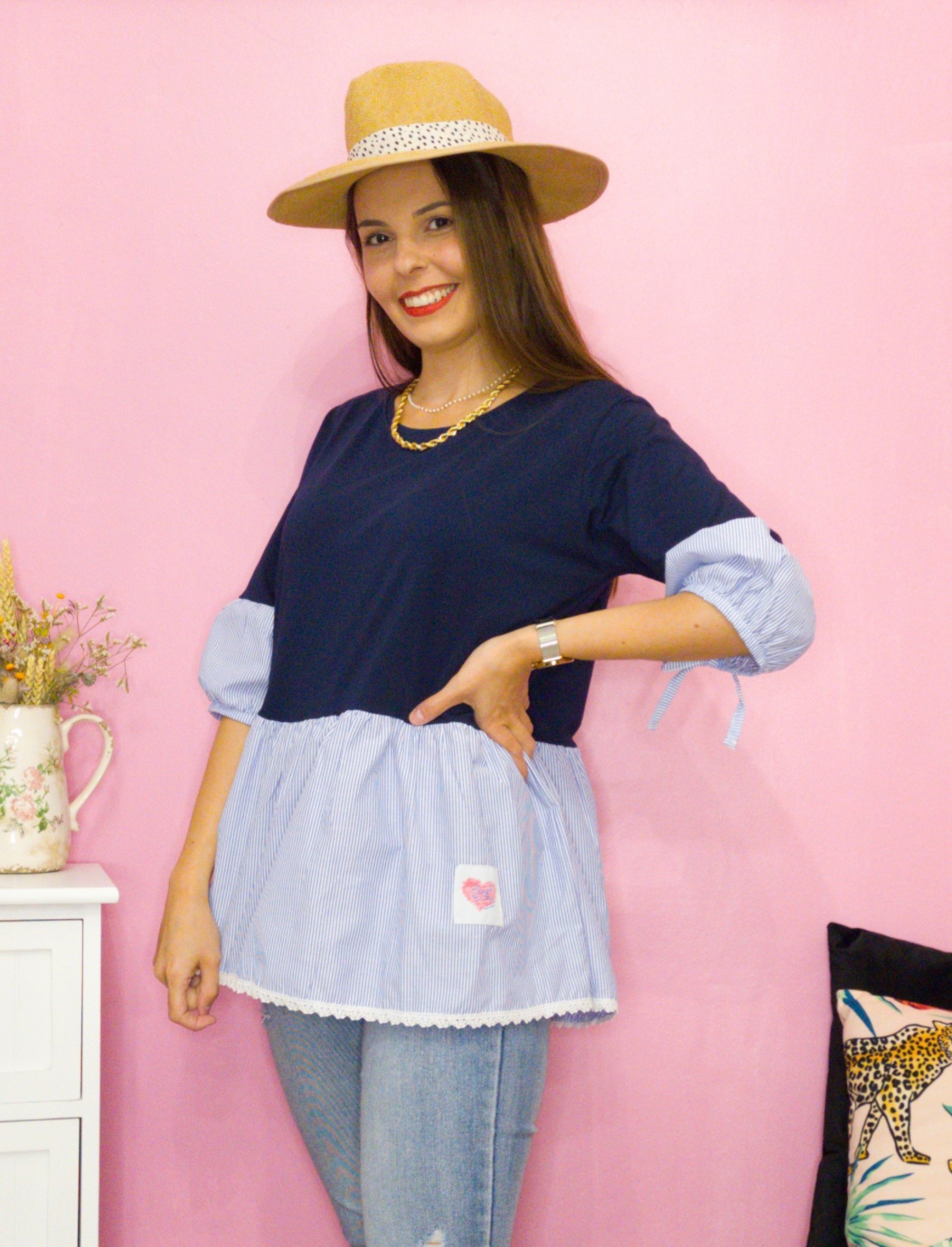 Mulher vestindo túnica azul bicolor com chapéu de palha posando contra parede rosa