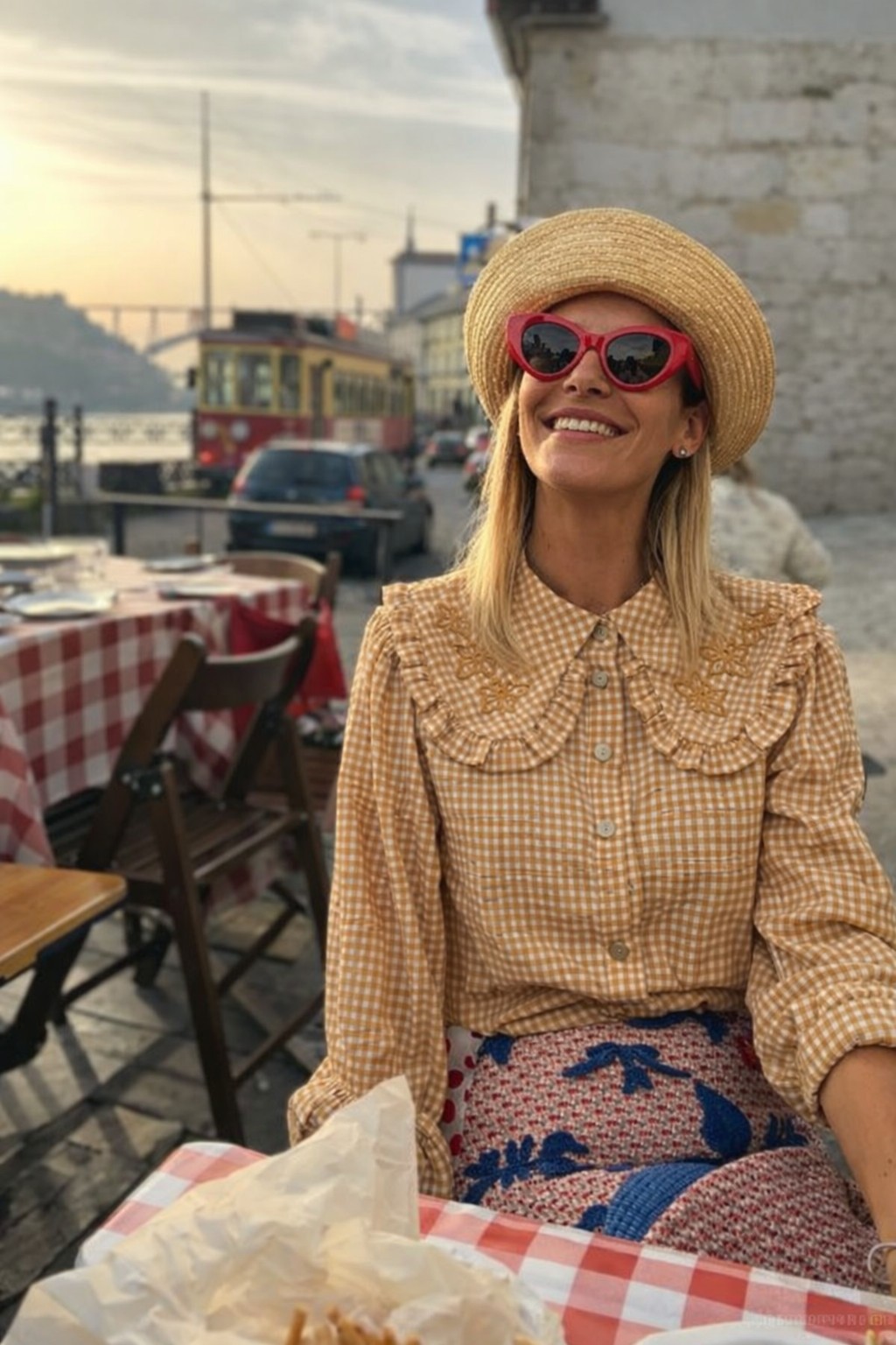 Mulher com chapéu de palha e óculos de sol vermelhos sentada numa mesa de restaurante com toalha xadrez