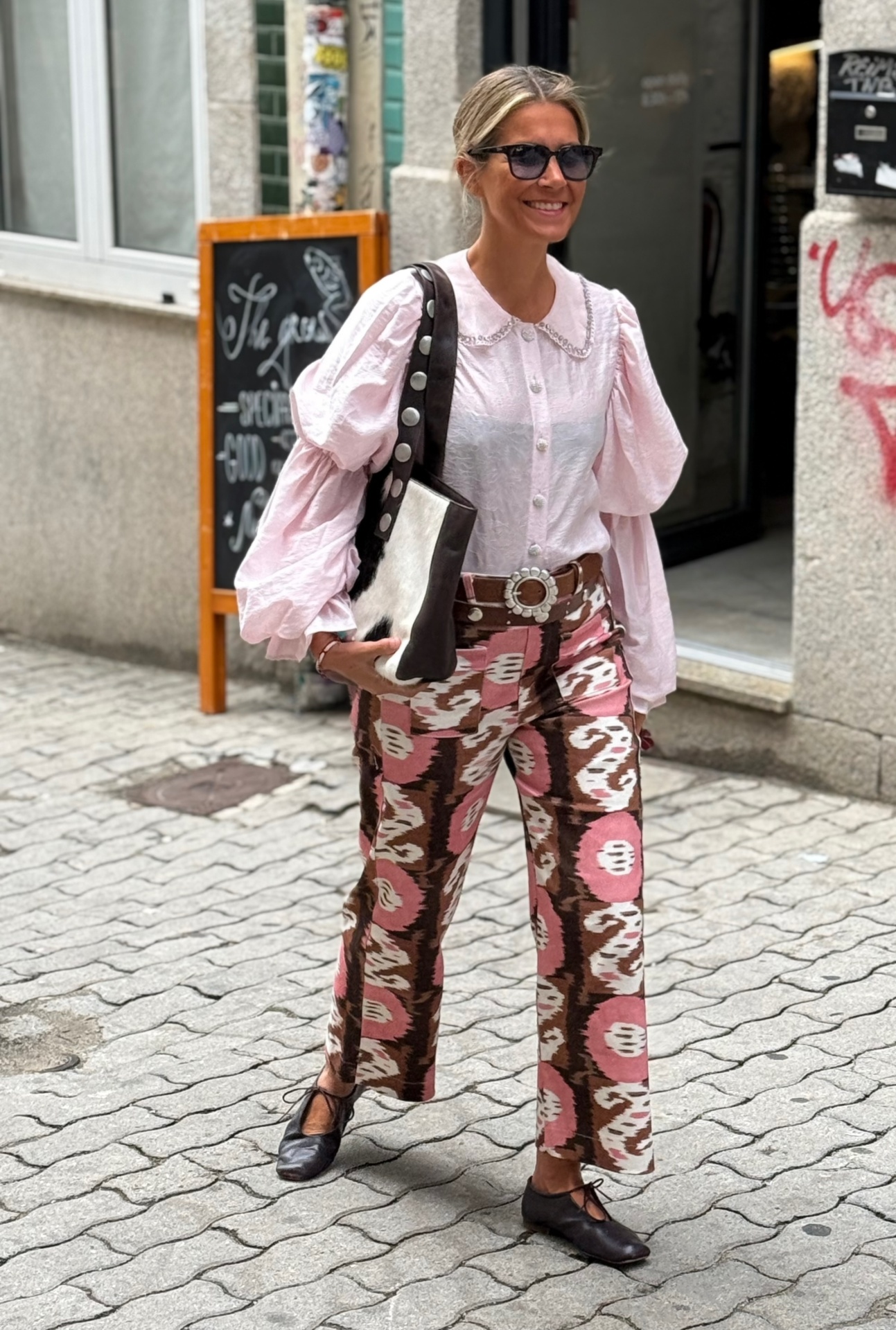 Mulher com camisa rosa, calças padrão rosa castanho, cinto castanho, sapatos pretos, óculos escuros e mala preta e branca.