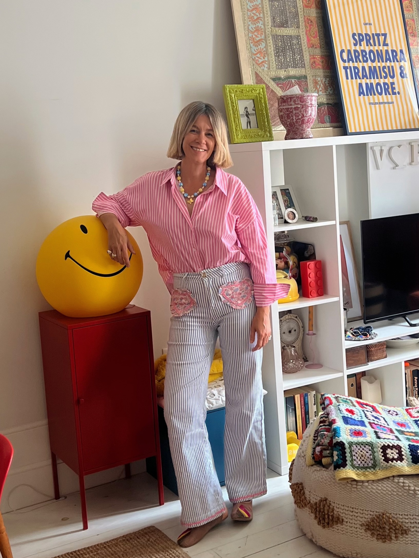 Mulher com camisa rosa às riscas e calças cinza às riscas, colar colorido, junto a baú vermelho com bola amarela sorridente em sala luminosa