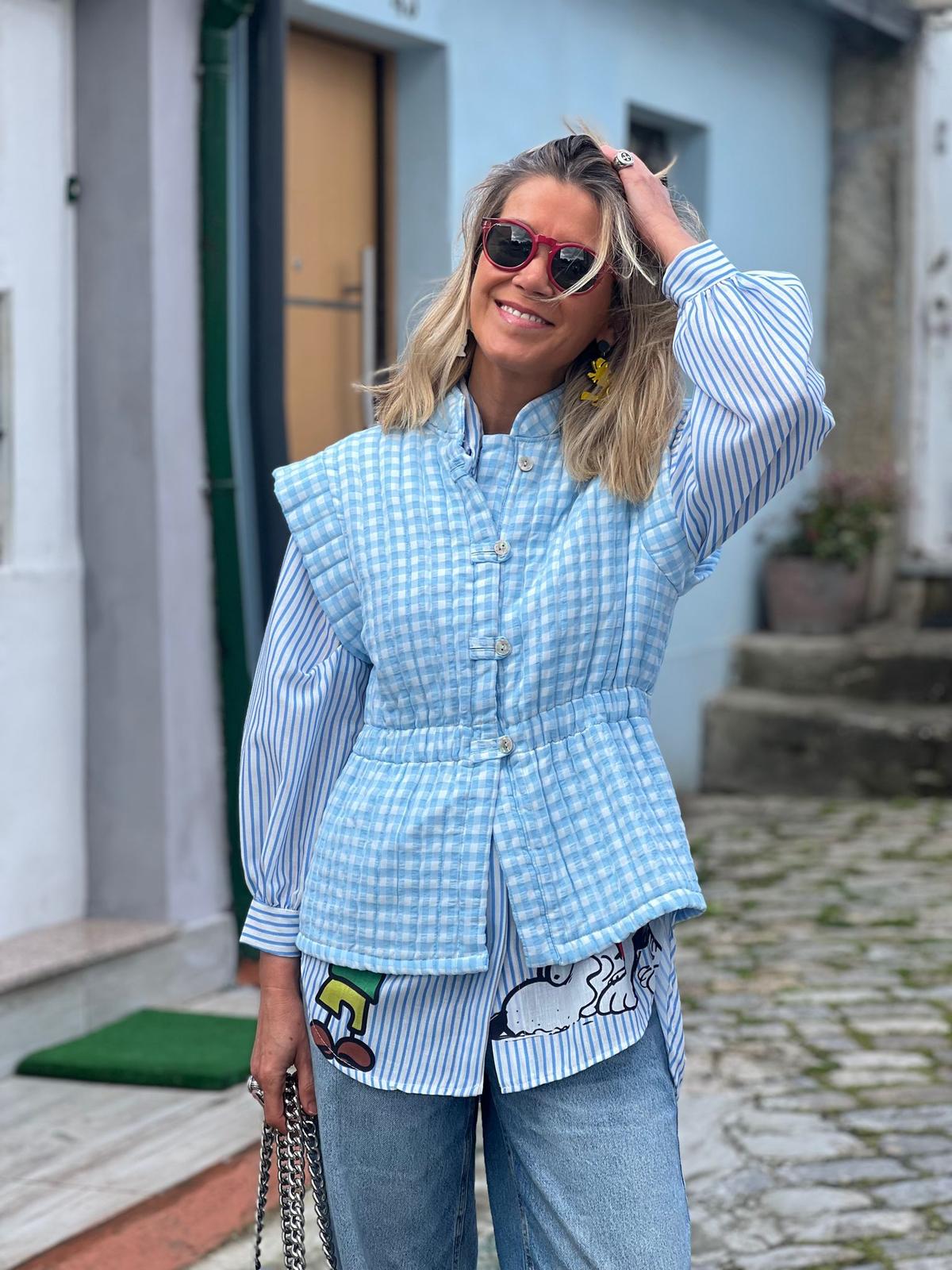 Mulher vestindo colete azul claro xadrez, camisa azul e branca riscada com estampa Snoopy, calças de ganga e óculos de sol vermelhos numa rua com calçada de pedra.