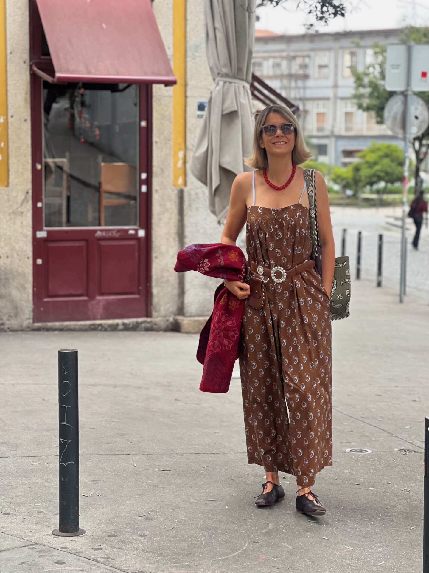 Mulher com vestido castanho floral, cinto branco, colar vermelho e óculos escuros caminhando na rua.