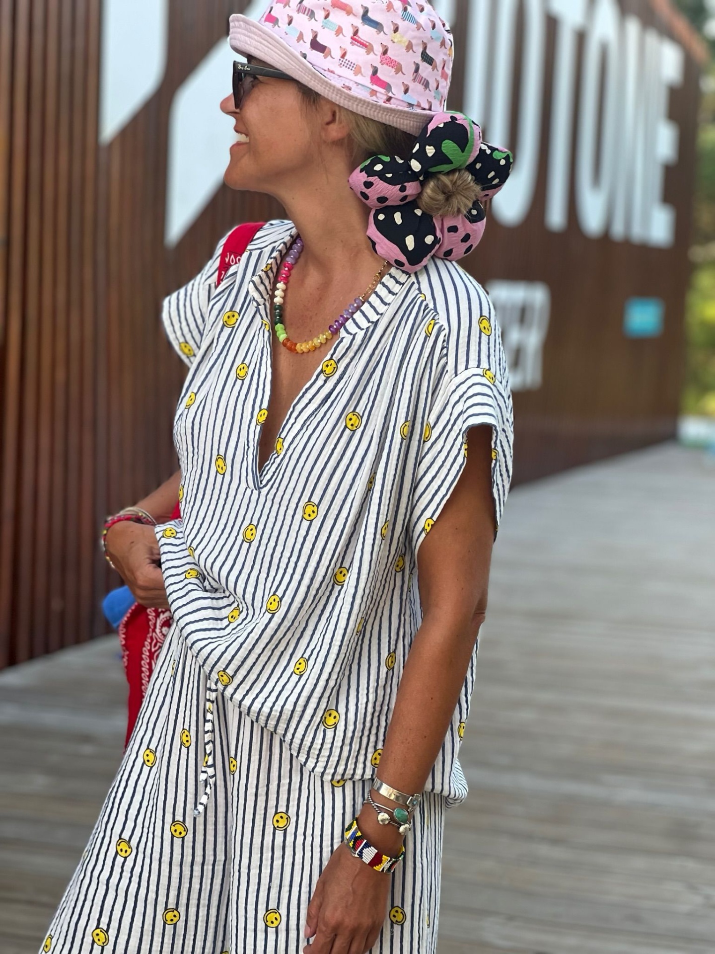 Pessoa com conjunto de roupa listrada com carinhas sorridentes e chapéu rosa com padrão de cães num passadiço de madeira