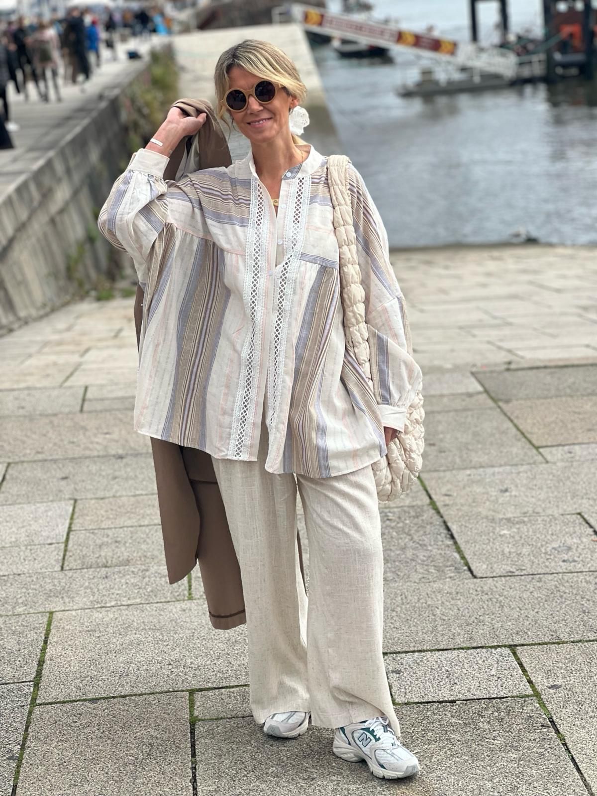 Mulher com blusa às riscas, calças largas creme, sapatilhas brancas e bolsa trançada junto a rio