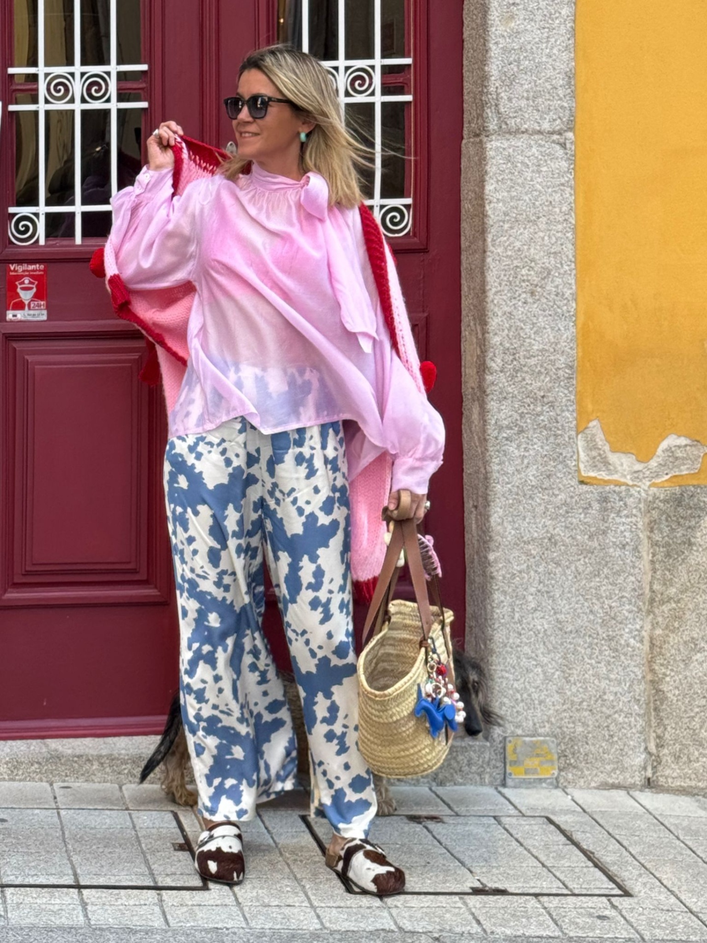 Mulher com blusa rosa, pantalones estampados azuis e brancos, sapatos brancos e marrons, bolsa de palha em frente a porta vermelha