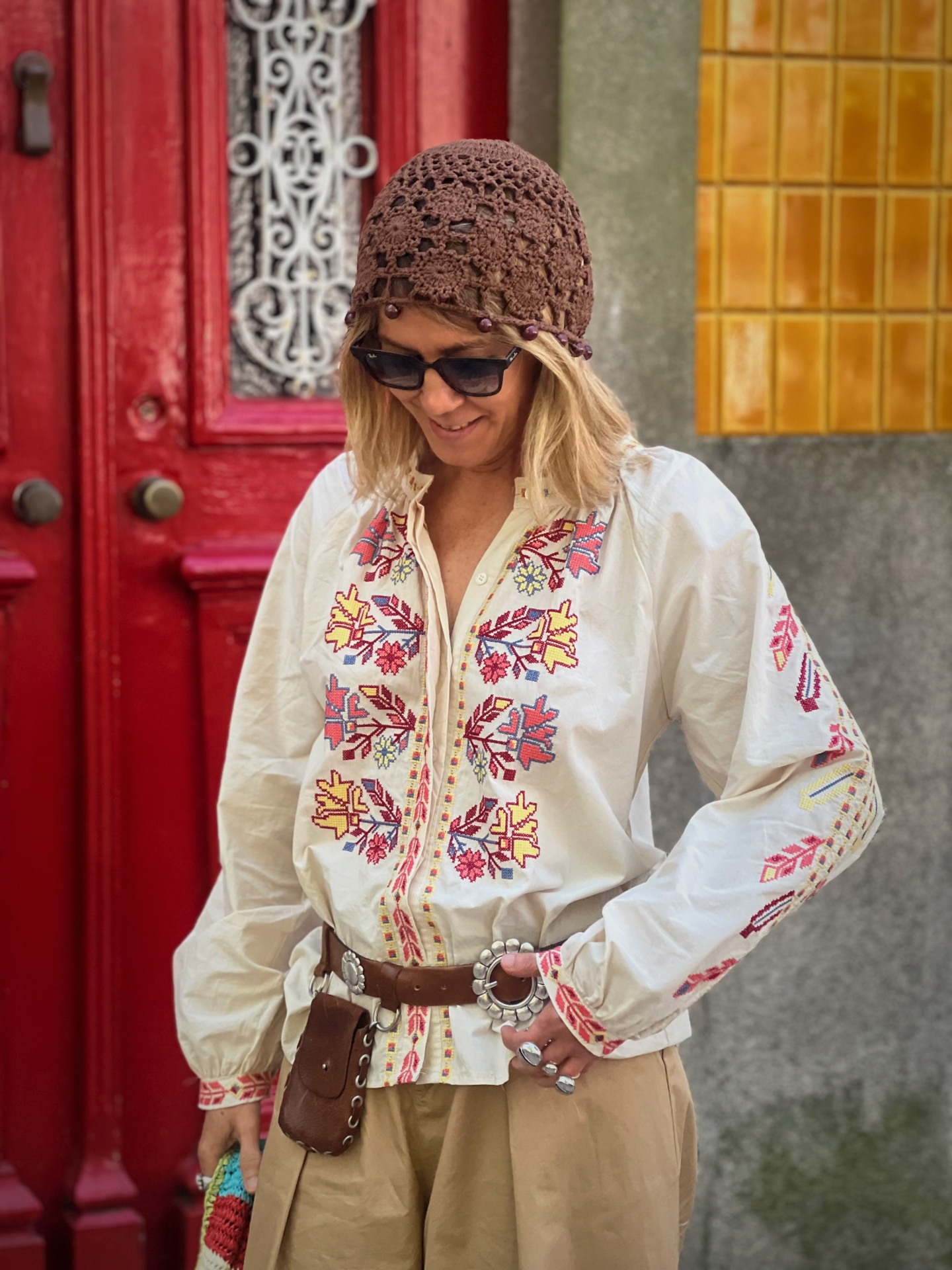 Blusa branca bordada colorida com cinto castanho, chapéu castanho de crochê e óculos escuros, mulher em ambiente urbano.