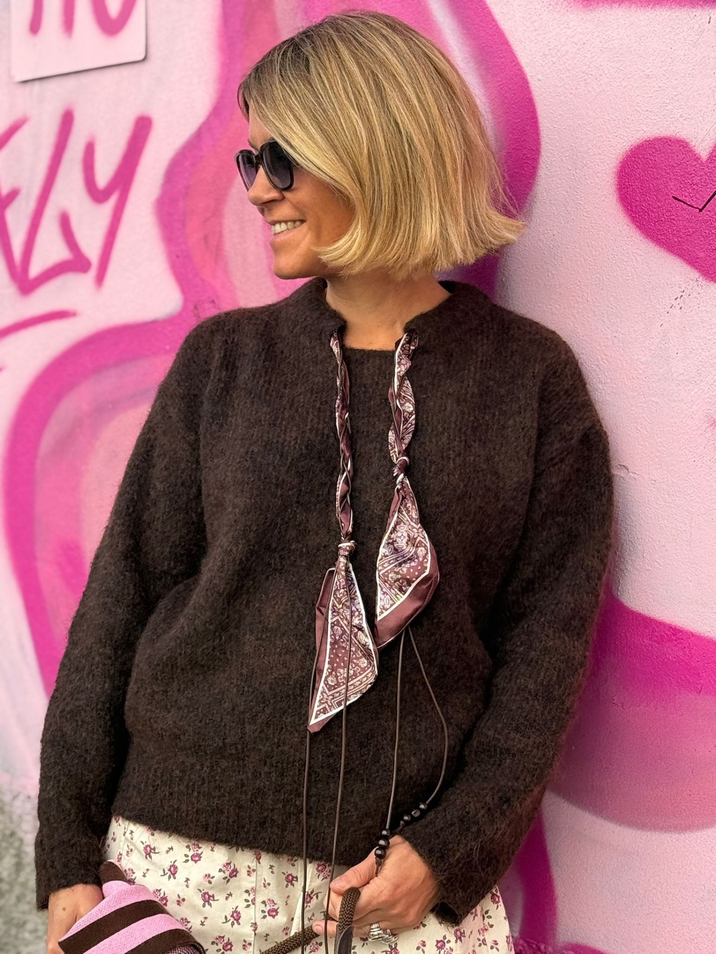 Mulher de cabelo loiro com camisola castanha felpuda junto a parede rosa com grafite.