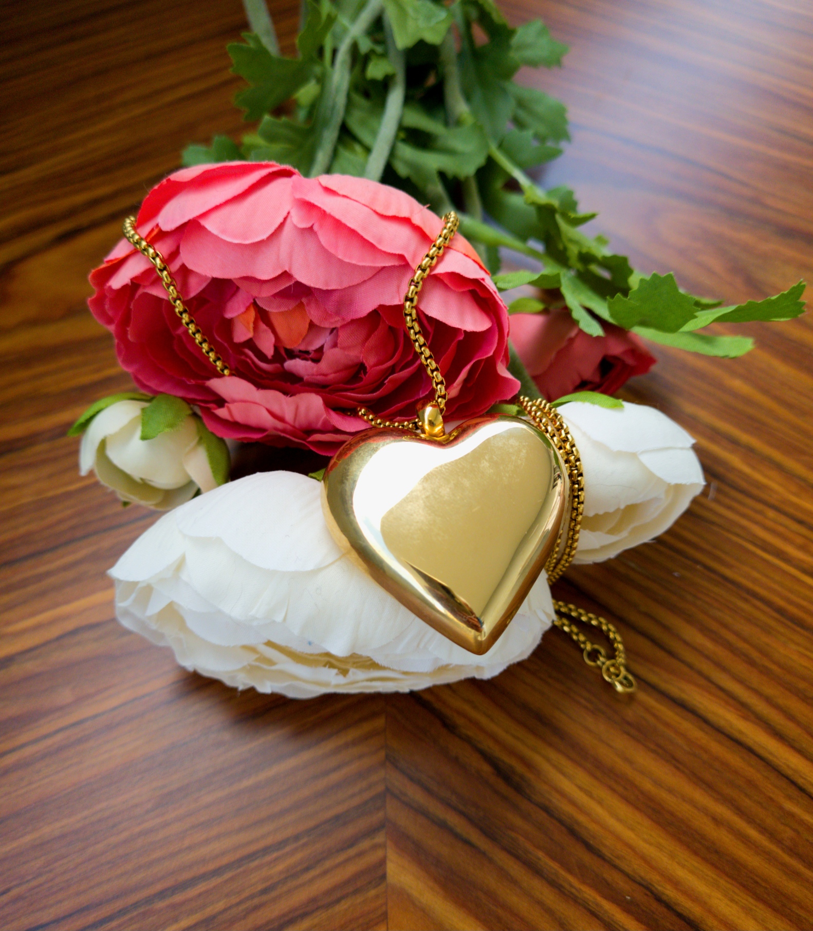 Colar dourado em forma de coração sobre flores branca e rosa em madeira clara