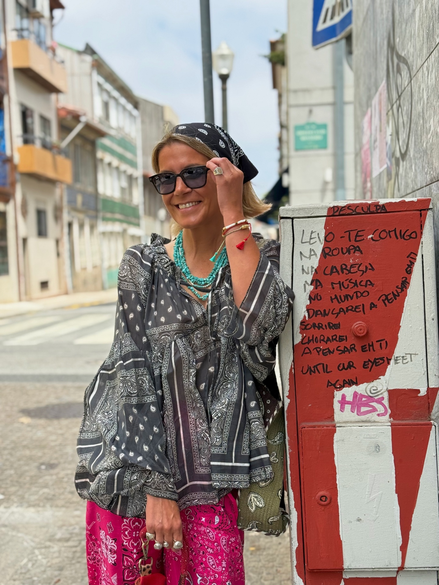 Mulher com blusa e lenço pretos com padrão paisley branco, calças rosa com padrão paisley, colares turquesa e pulseiras, junto a poste com graffiti e texto em rua urbana