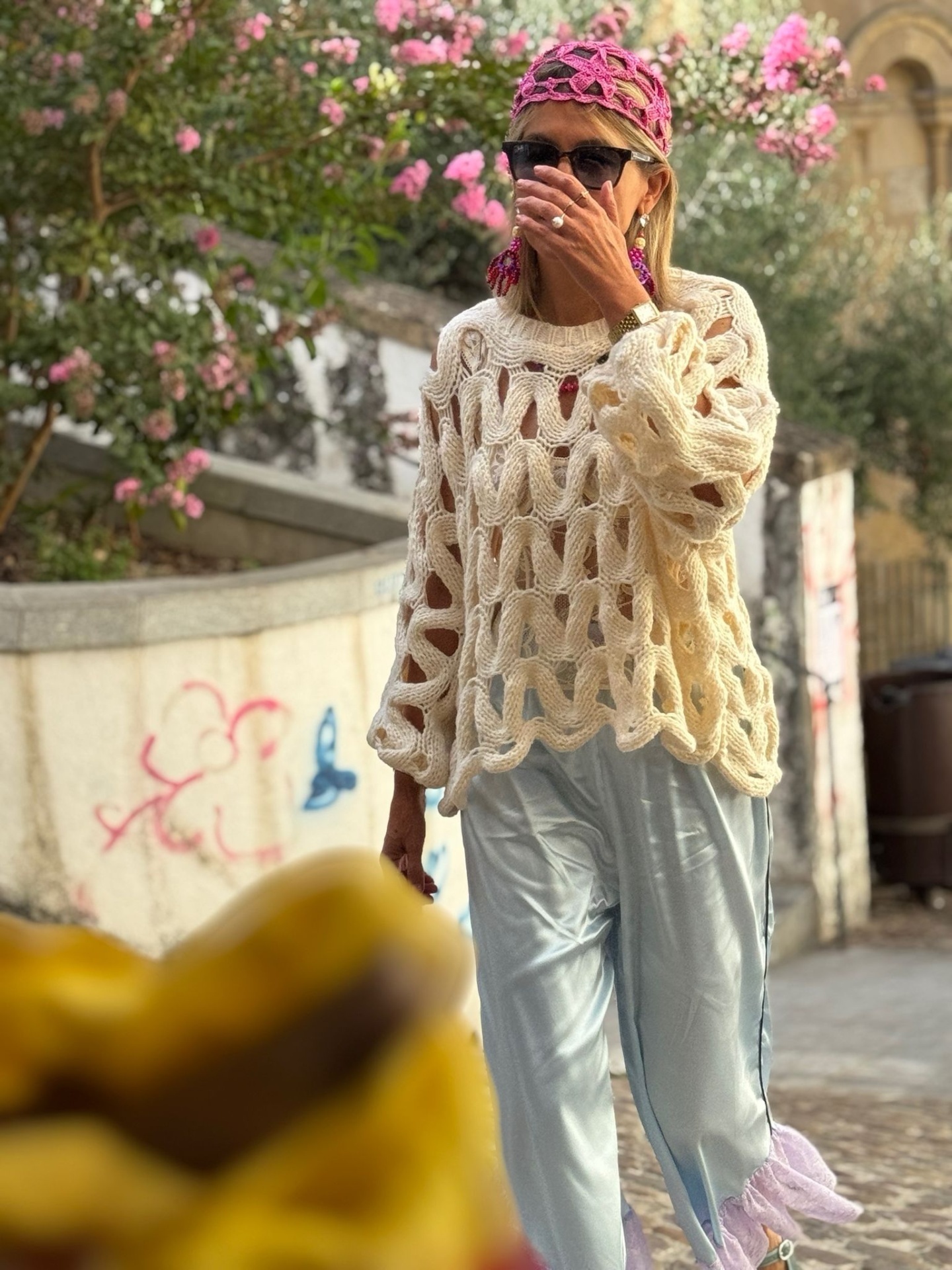Pessoa com camisola de malha creme de laços grandes e calças cinza numa rua com flores cor-de-rosa.