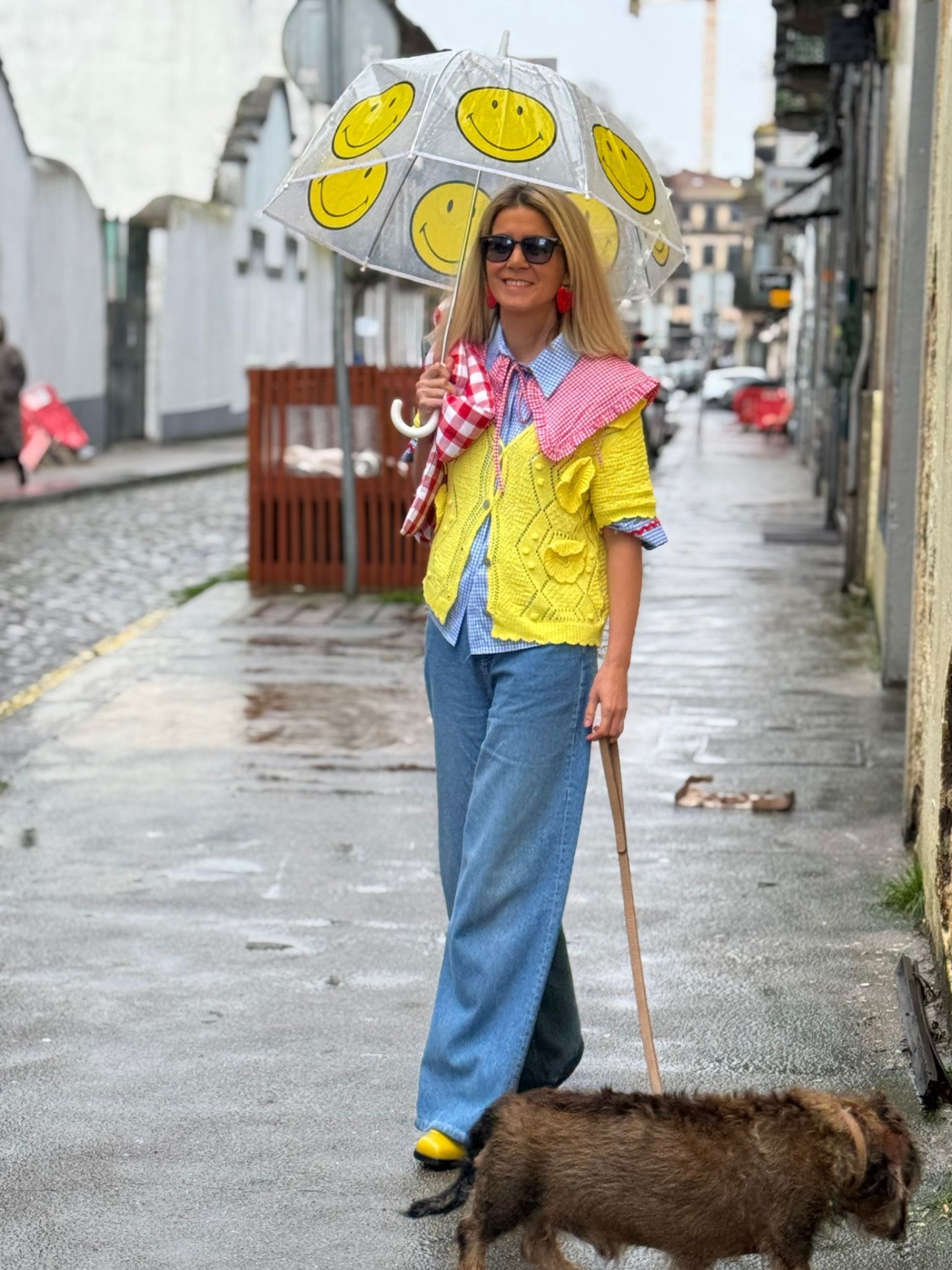 Mulher com guarda-chuva transparente com carinhas sorridentes, colete amarelo e calças azuis em rua molhada