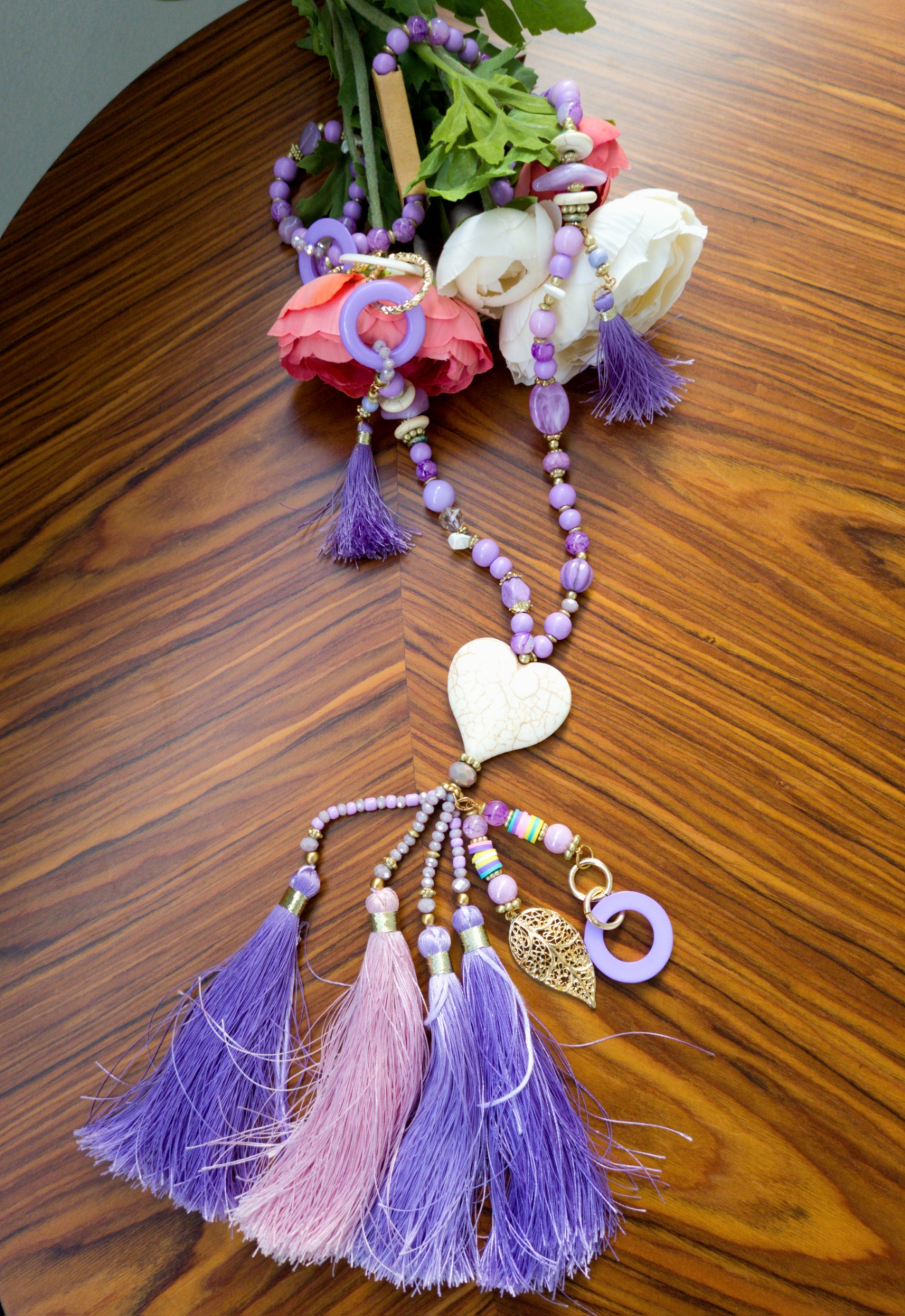 Colar com contas lilases, pingentes em forma de coração, borlas coloridas sobre mesa de madeira e flores artificiais