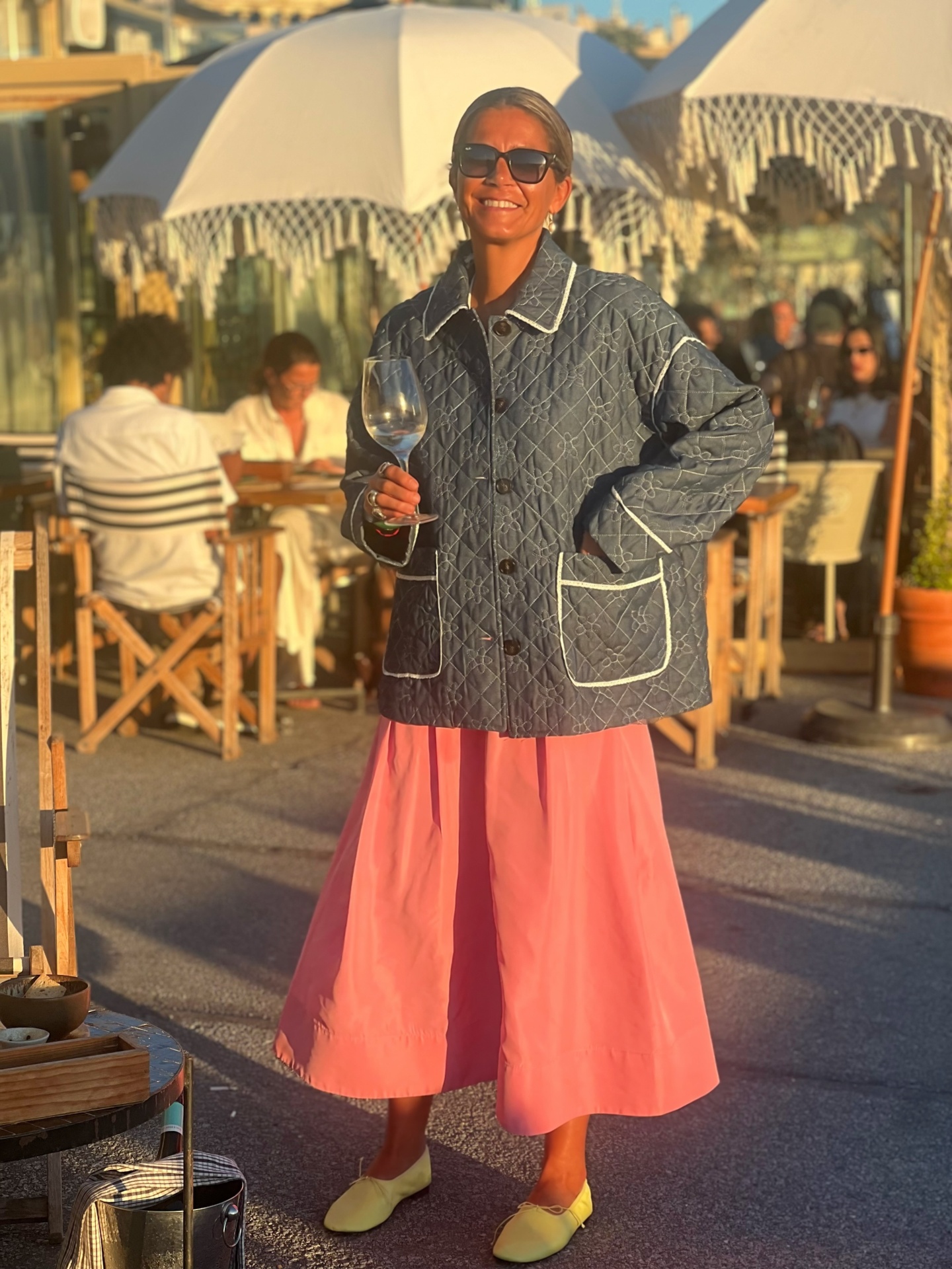 Mulher com casaco acolchoado cinzento, vestido rosa e sapatos amarelos segura copo em esplanada com guarda-sóis brancos