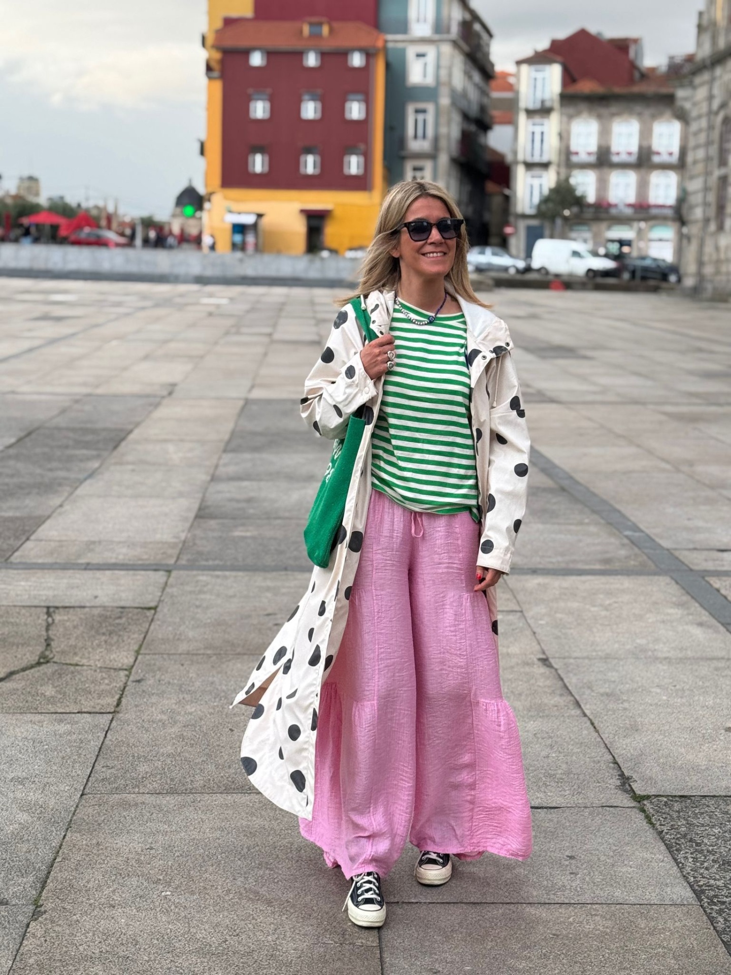 Mulher com casaco branco de bolas pretas, blusa verde às riscas, calças rosa largas e sapatilhas num espaço urbano