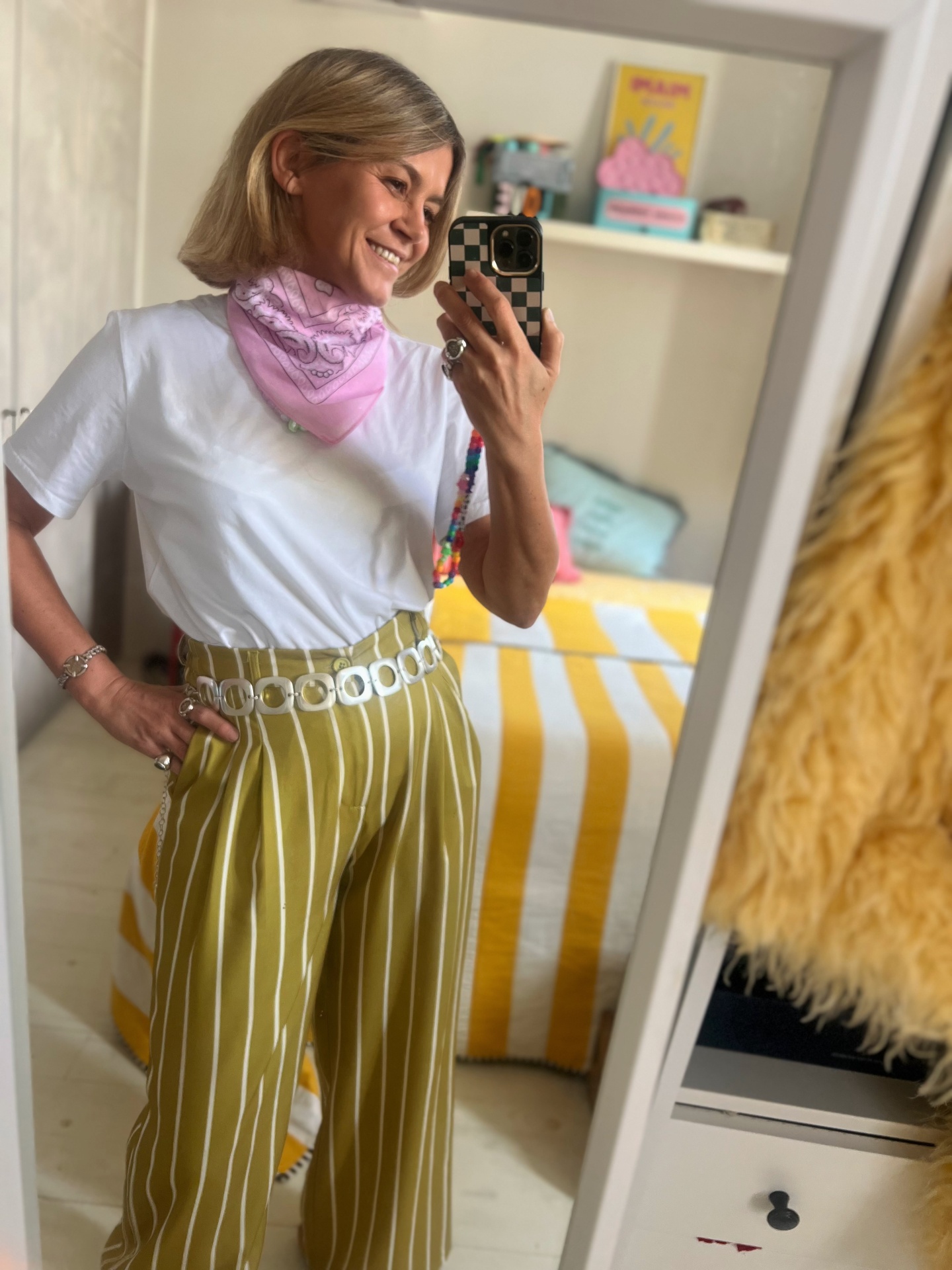 Pessoa a sorrir com roupa branca, lenço cor-de-rosa e calças largas riscas verticais verdes e brancas.