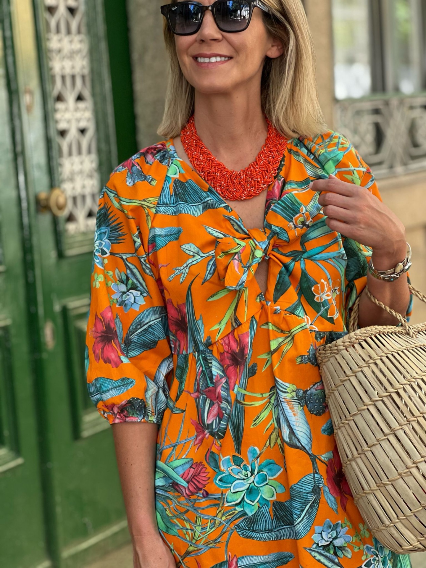 Mulher com vestido floral laranja, colar vermelho, bolsa de palha, óculos de sol e porta verde com ferragens em fundo