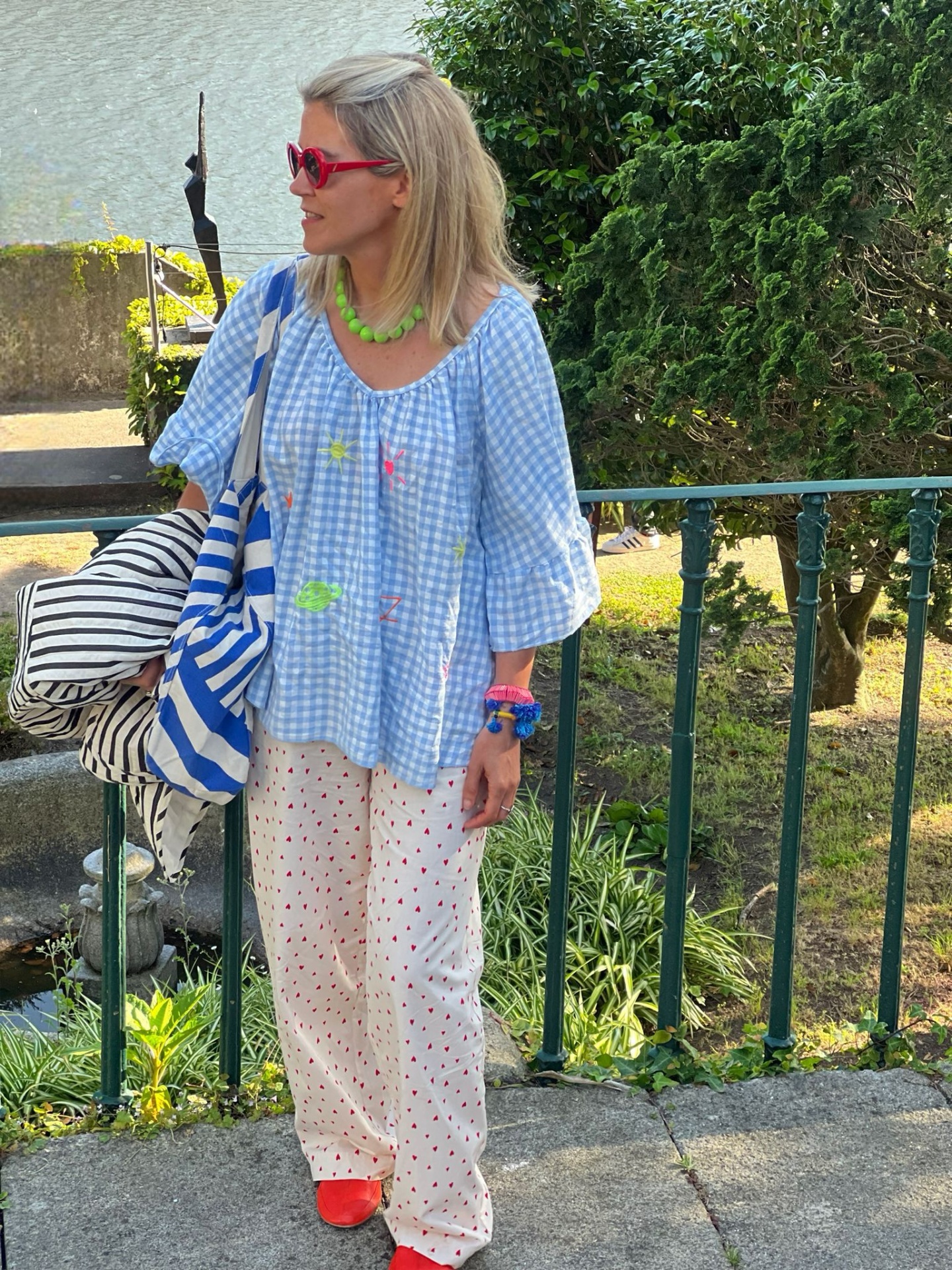 Mulher com túnica azul clara xadrez e calças brancas com corações vermelhos com acessórios coloridos e sacos listrados