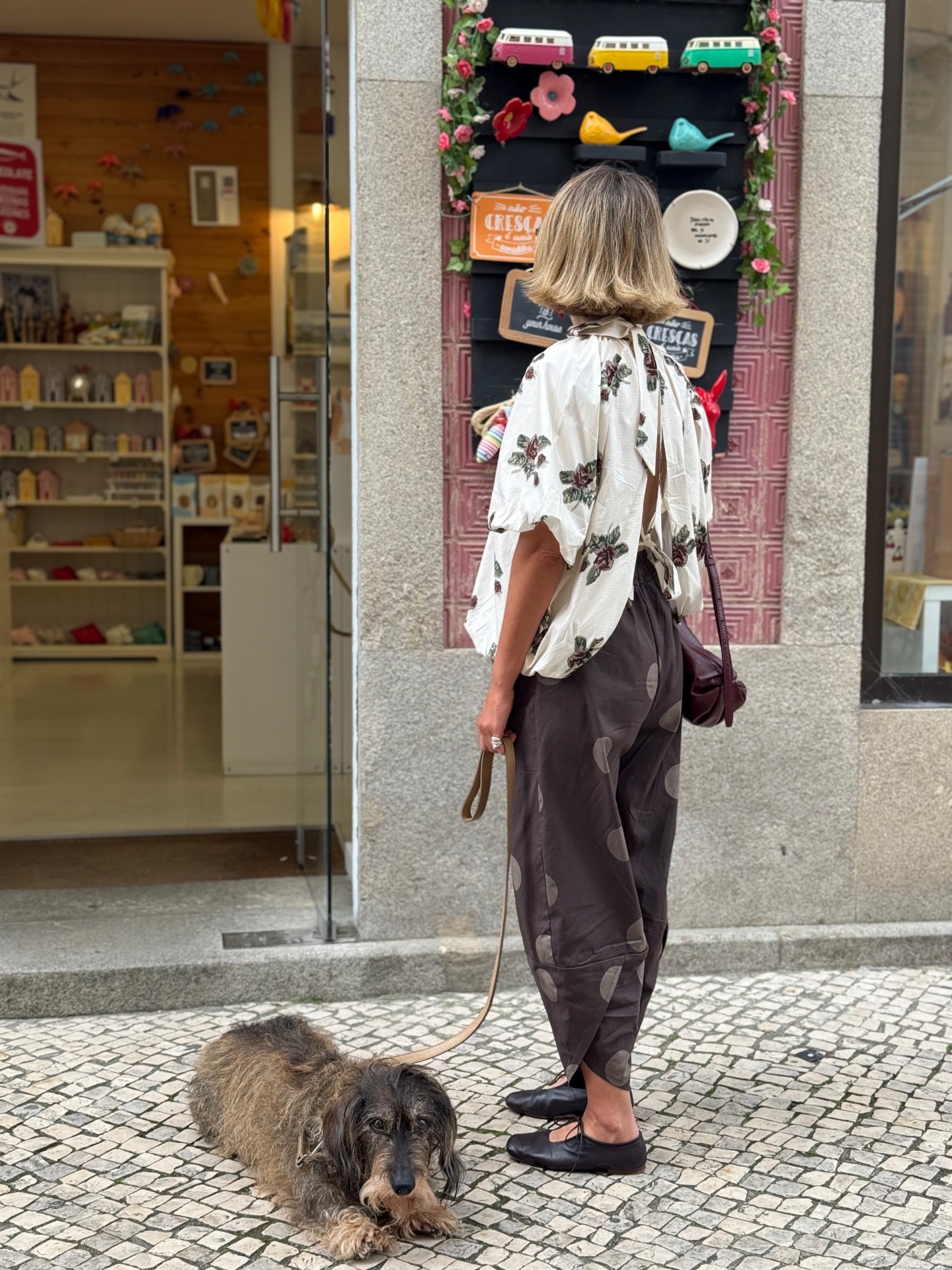 Mulher de costas com blusa floral e calças castanhas com cão de trela no chão em frente a loja decorada.