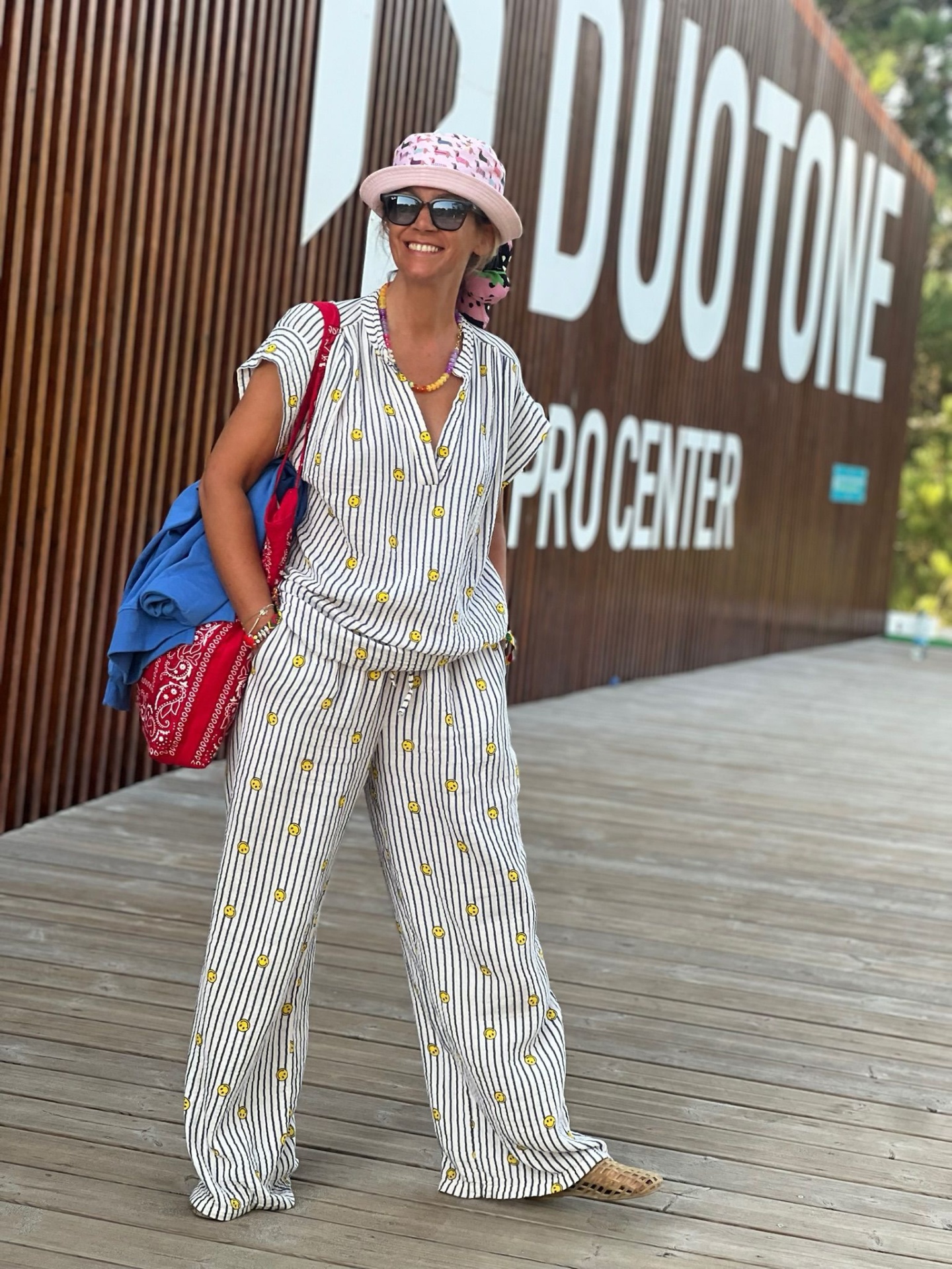 Mulher com roupa clara de riscas e flores, chapéu rosa, óculos, mochila azul e saco vermelho em passadeira de madeira junto a painel DUOTONE PRO CENTER.