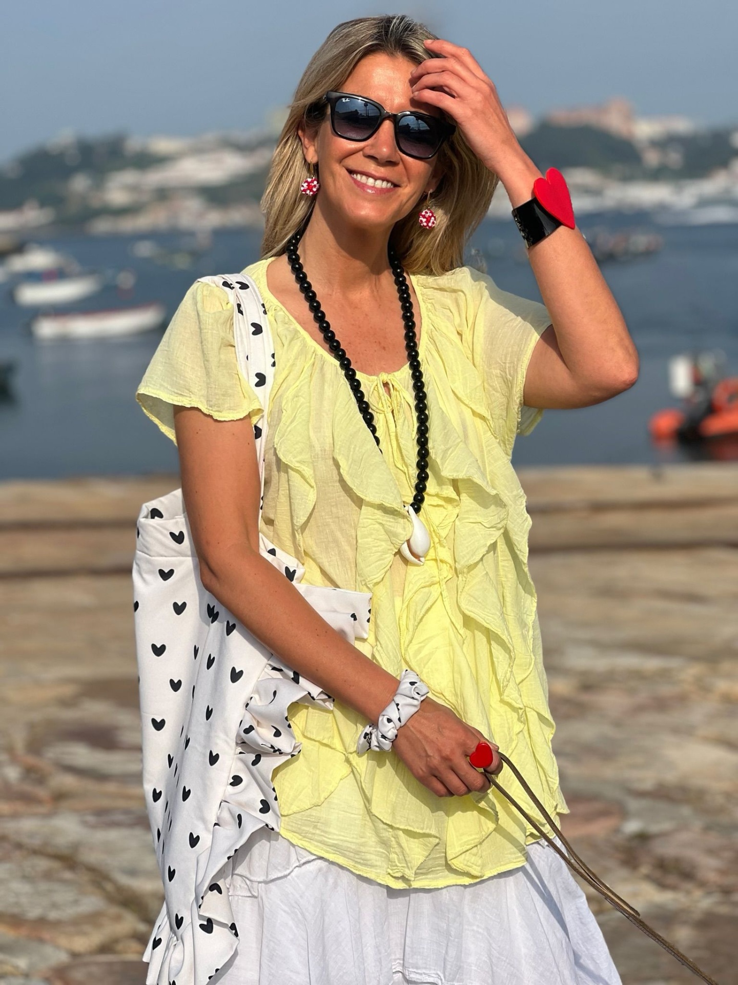 Mulher com blusa amarela e saia branca junto ao mar, acessórios coloridos e bolsa branca com corações pretos.